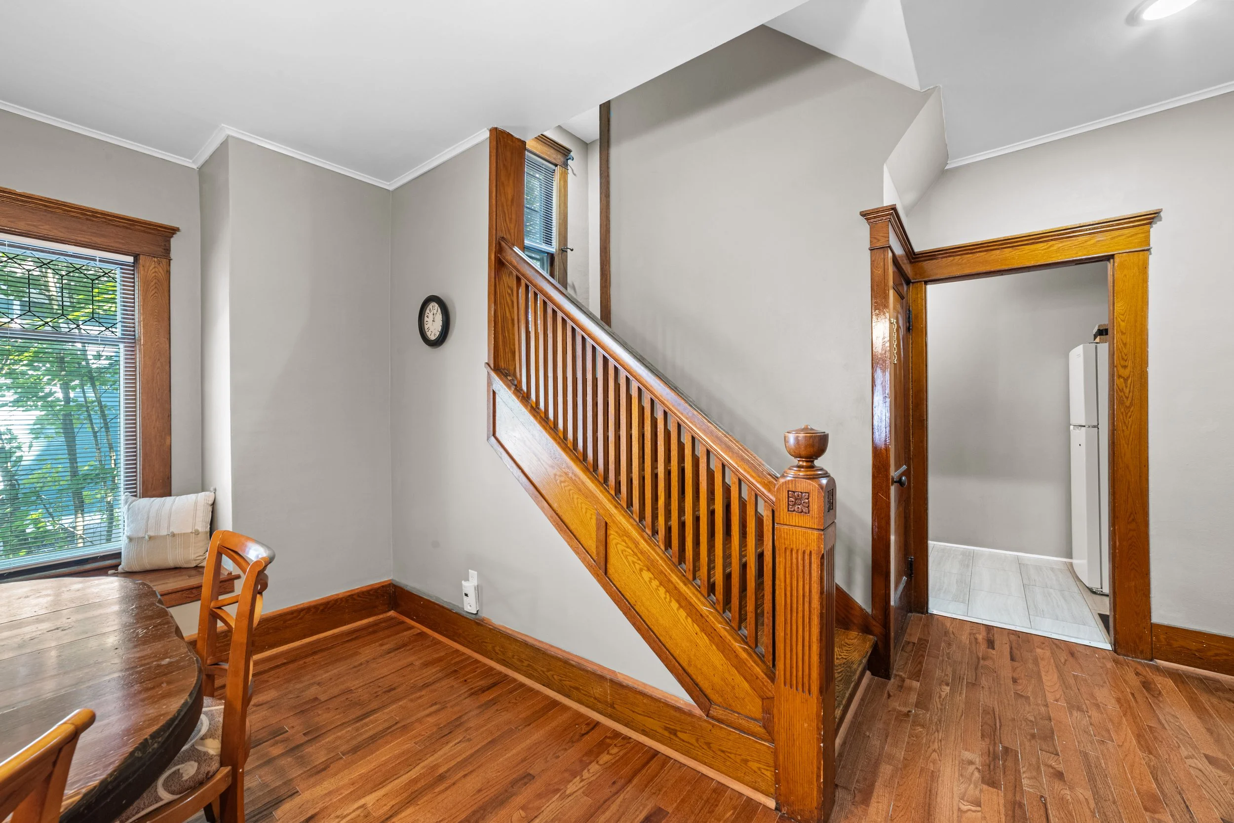 Interior view of a home showing a staircase with wooden railing, a clock on the wall, a window with blinds, a doorway leading to a laundry area with a white refrigerator, and hardwood floors.