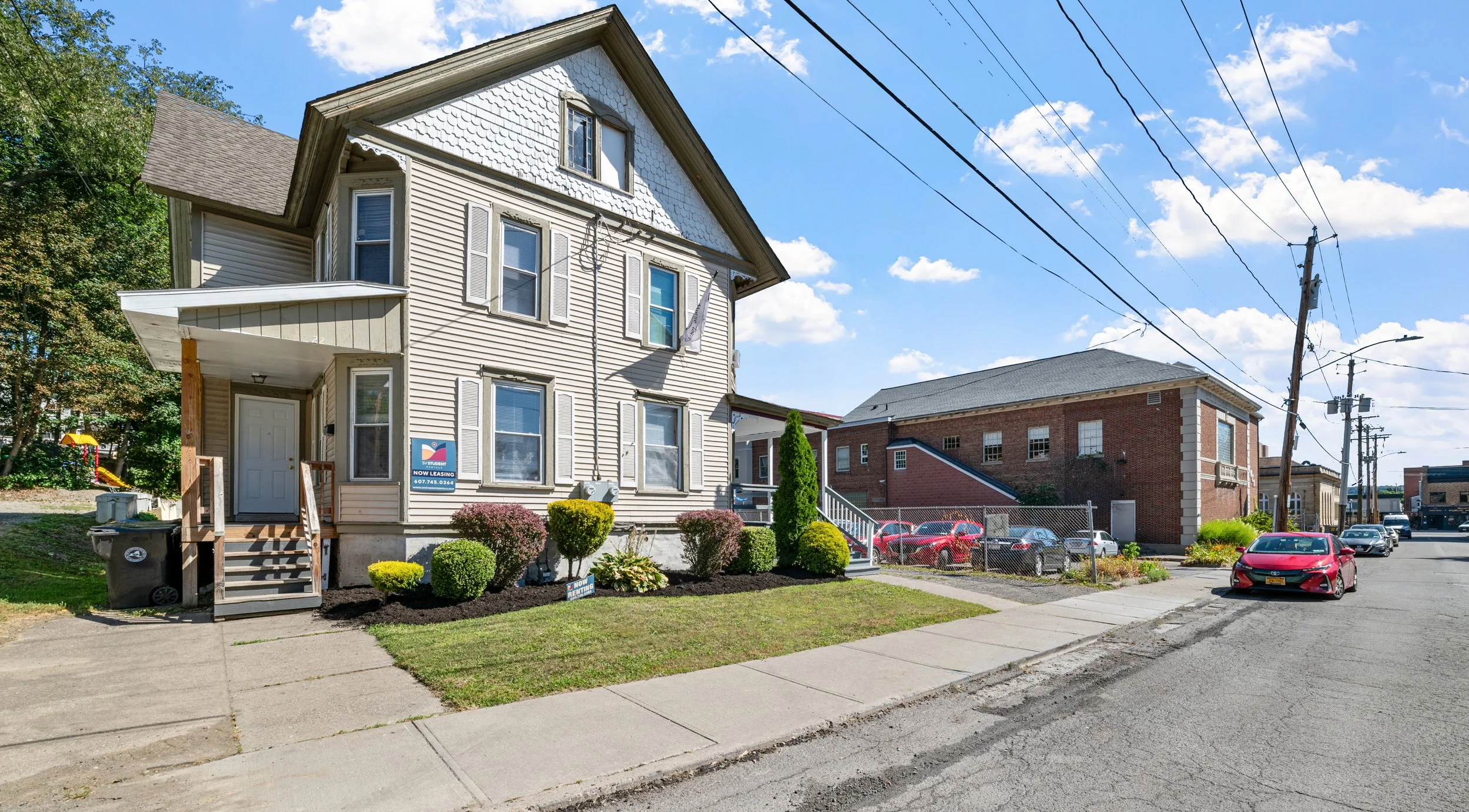 A multi-story house with beige siding, front garden with shrubs and flowers, and a small staircase leading to the front door under a covered porch. There are signs indicating it is available for rent.