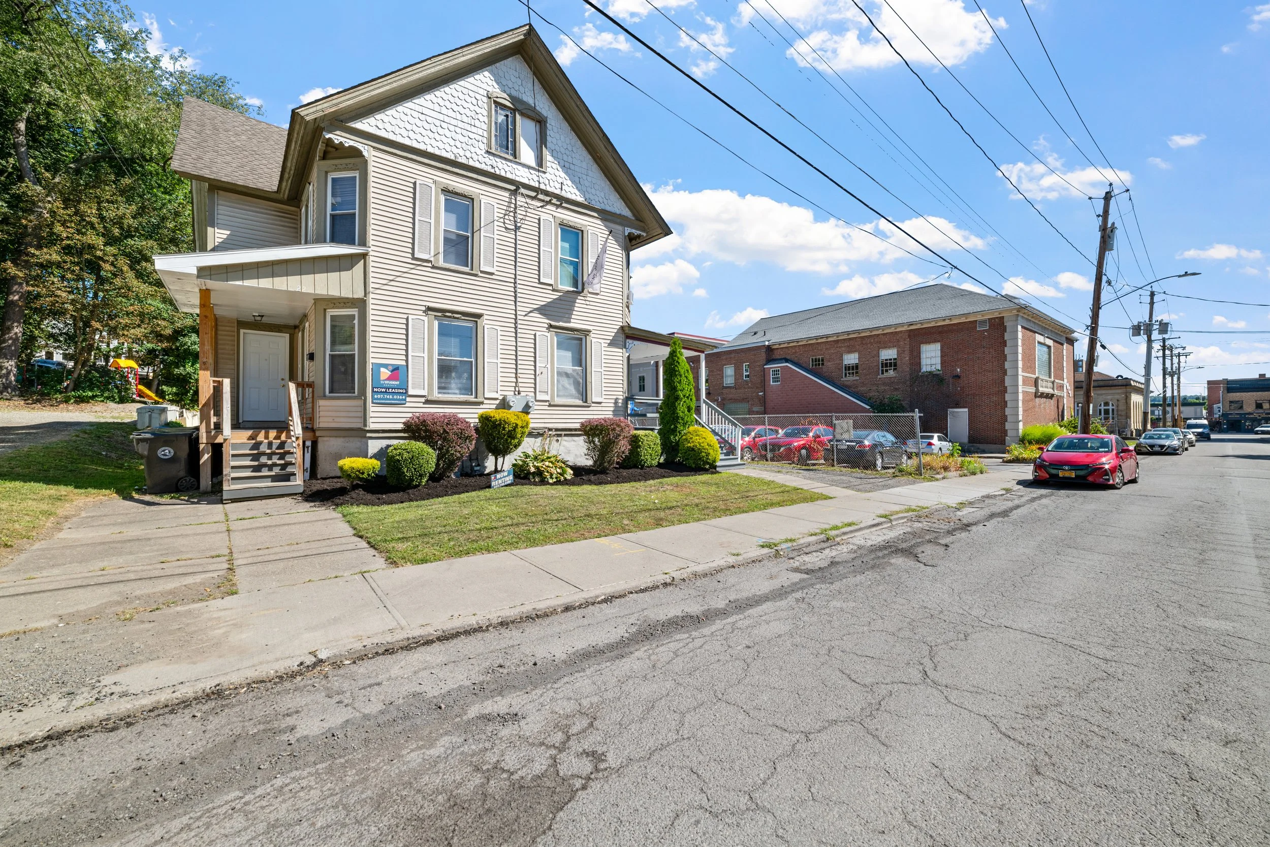 A residential street with a white, three-story house featuring beige shutters and a small front yard with bushes. There is a sign indicating the house is for lease. The street has parked cars and power lines overhead, with a blue sky and some clouds.
