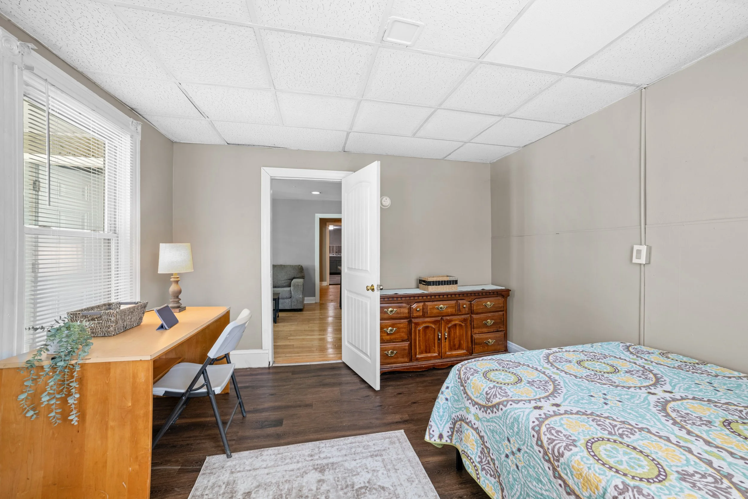 A bedroom with a bed covered in a colorful patterned quilt, a wooden dresser, a small desk with a lamp and a potted plant, a window with blinds, and an open door leading to a living area with hardwood floors and a gray couch.