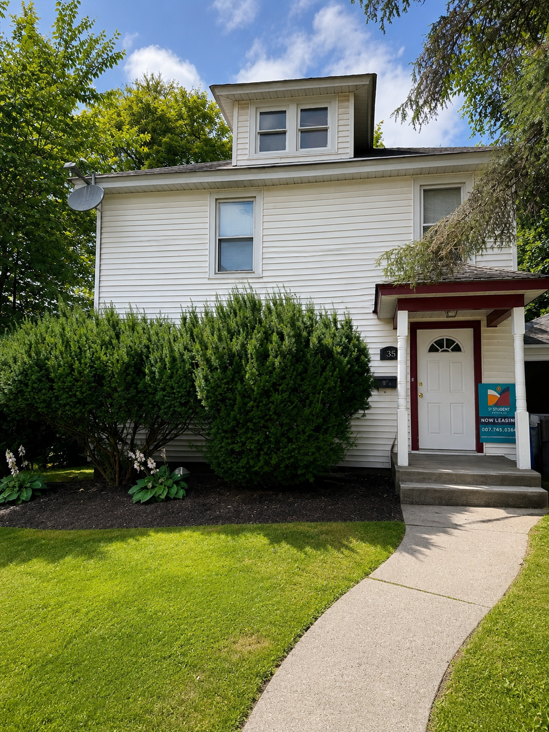 A two-story house with beige siding, a small front porch, and a gabled roof. There are two bushes in front of the house and a large tree on the right side. A satellite dish is attached to the left side of the house, and a black car is parked in the driveway on the right. The house has a sign indicating it is for student rental.