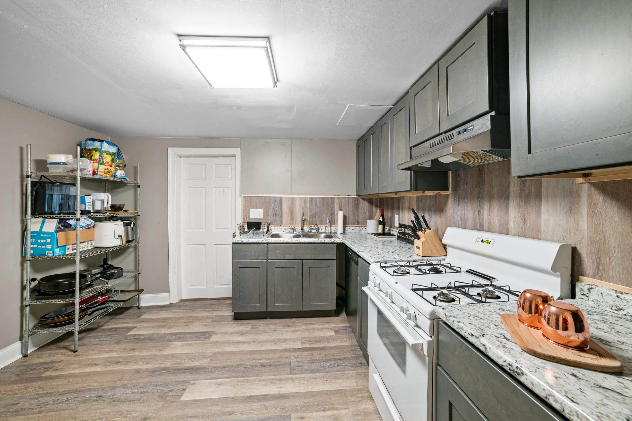 Kitchen with grey cabinets, white stove, granite countertops, wood flooring, and a metal shelving unit with kitchen supplies.