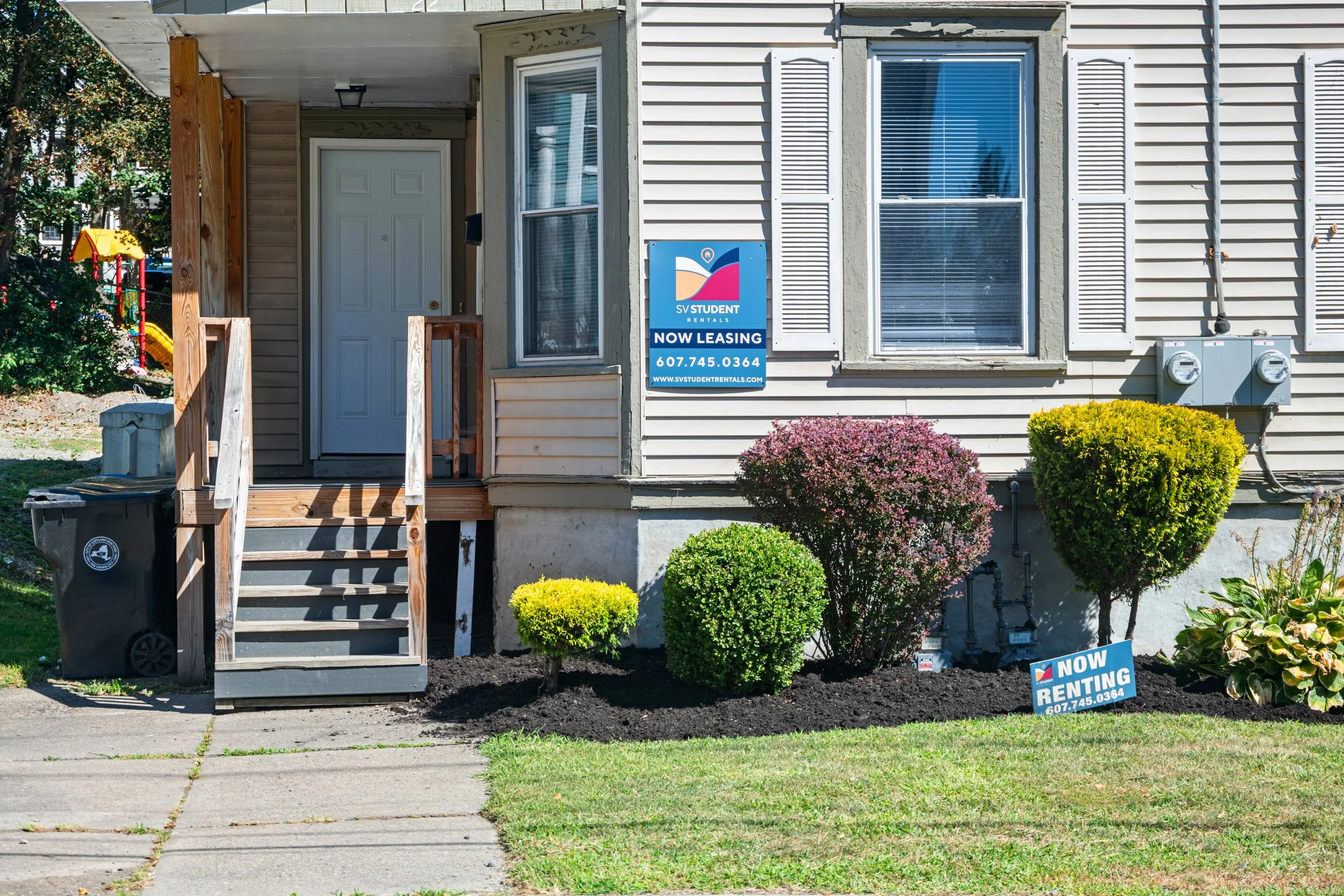 Front of a house with a small porch, staircase, and landscaped front yard with bushes. Two signs advertise the property for lease and rent, with contact information. There are utility boxes on the exterior wall.