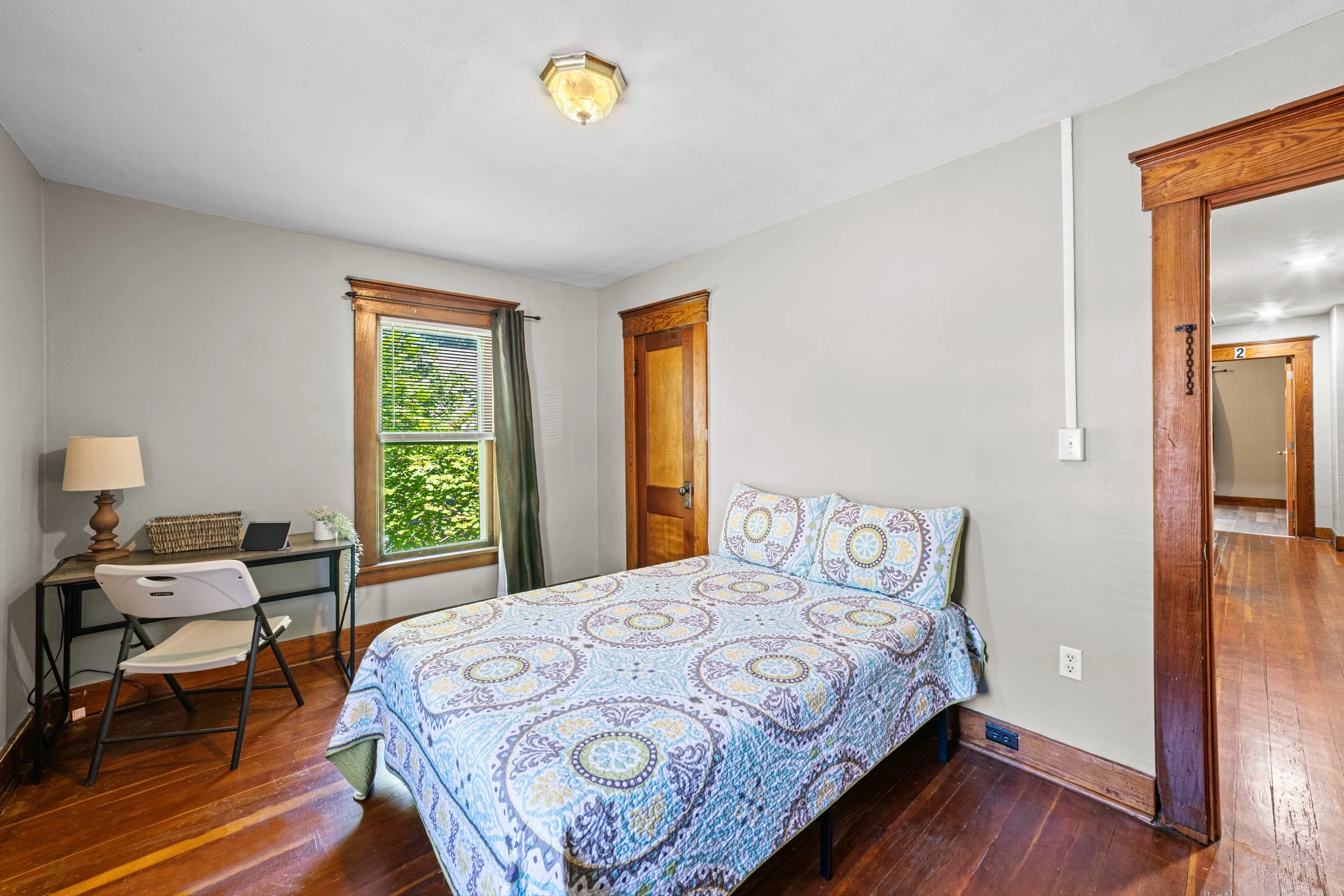 A bedroom with a wooden floor, a bed with a patterned bedspread and pillows, a small desk with a lamp and a chair near a window with green curtains, and a wooden door.