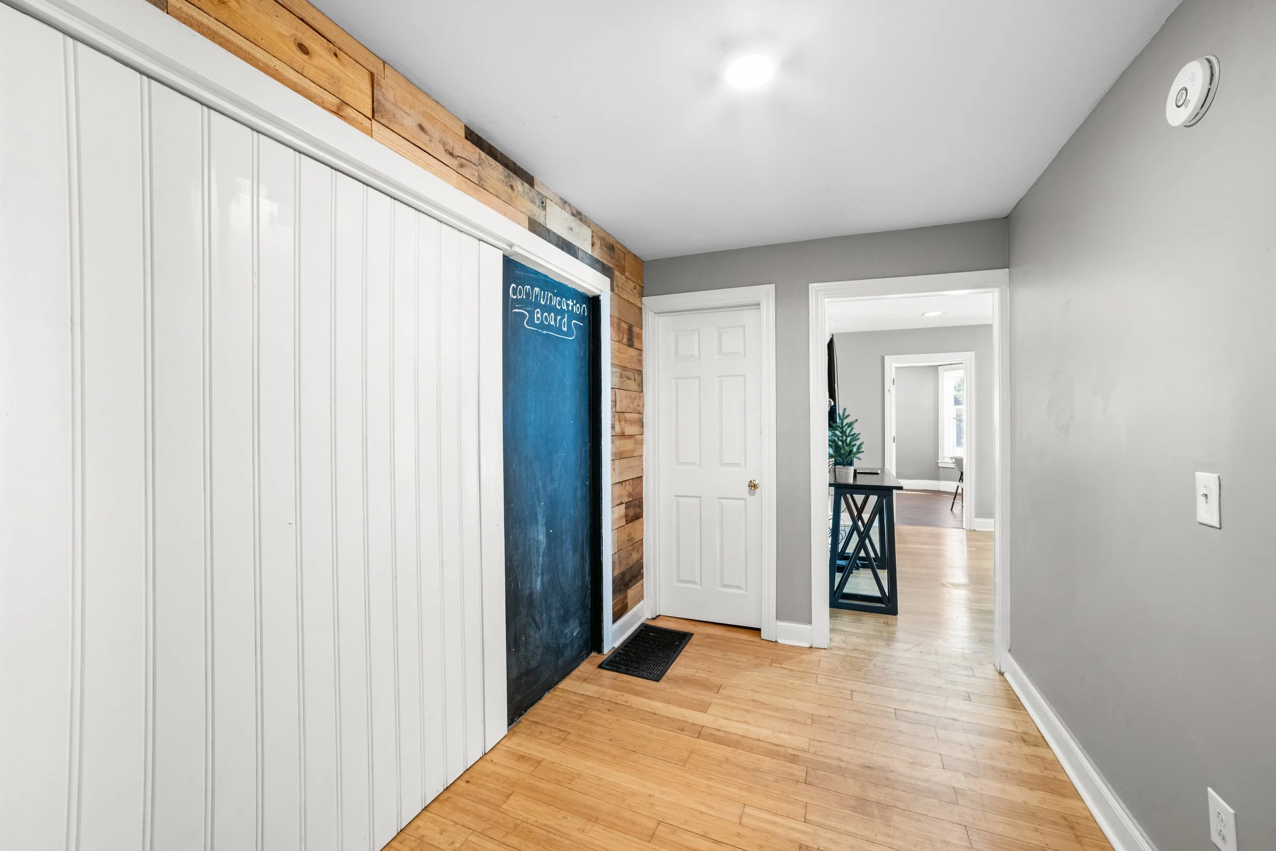 Interior hallway with a gray wall, a small table with plants, a white door, and a chalkboard labeled 'Communication Board' on the wall.