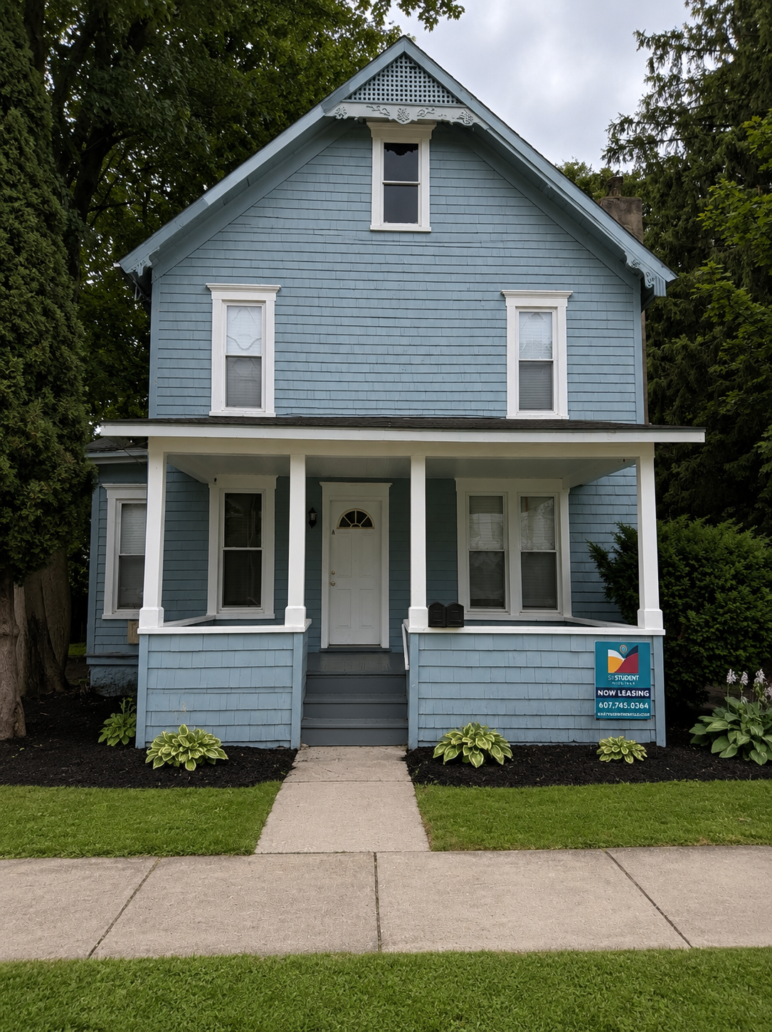 A two-story blue house with white trim and a porch, surrounded by trees, with a sign in front advertising student rentals and phone number.