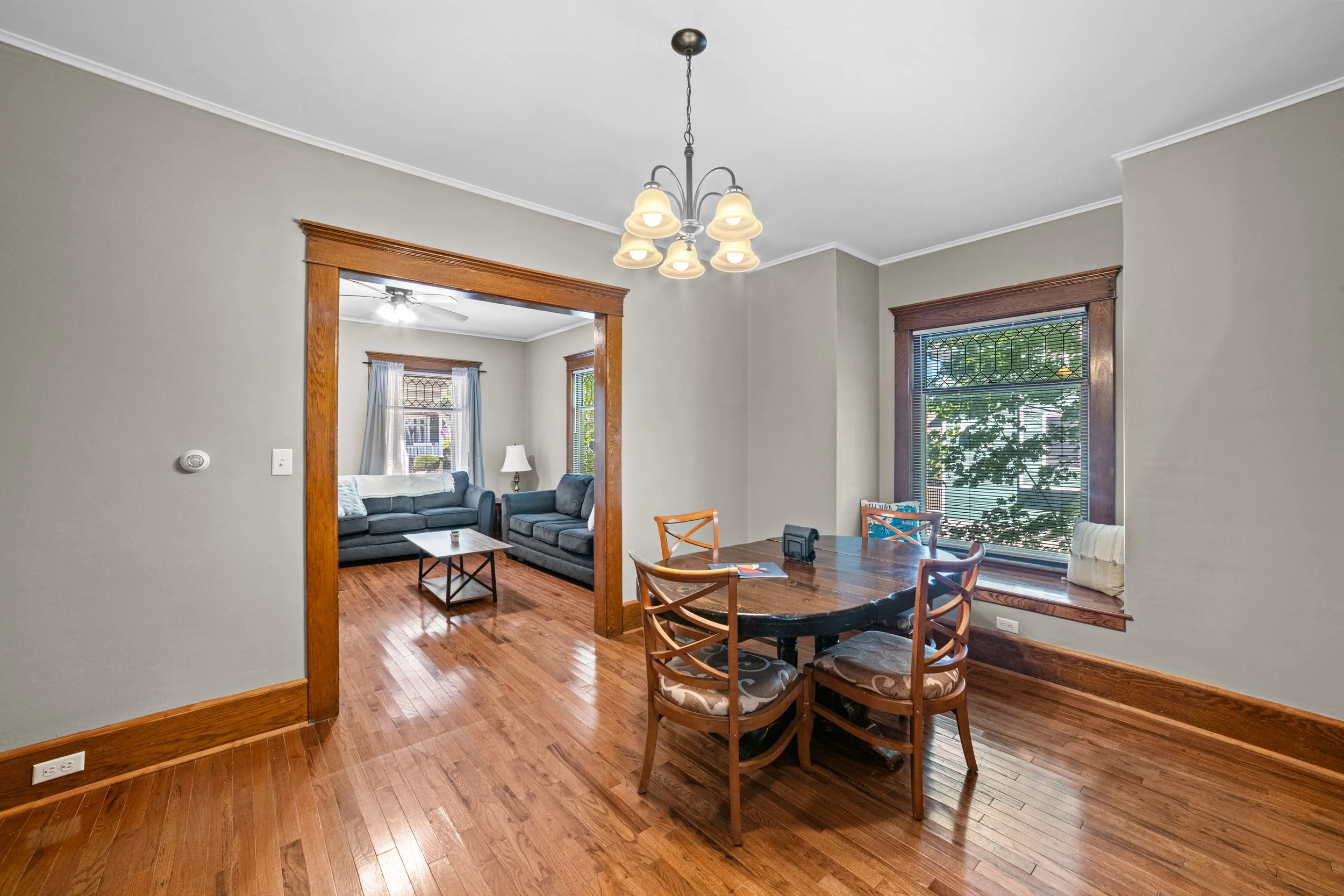 Dining room with wooden floor, a dark wooden oval table with four matching chairs, window with blinds, and a chandelier overhead. Connected to a living room with two gray sofas, a coffee table, and a window with curtains.