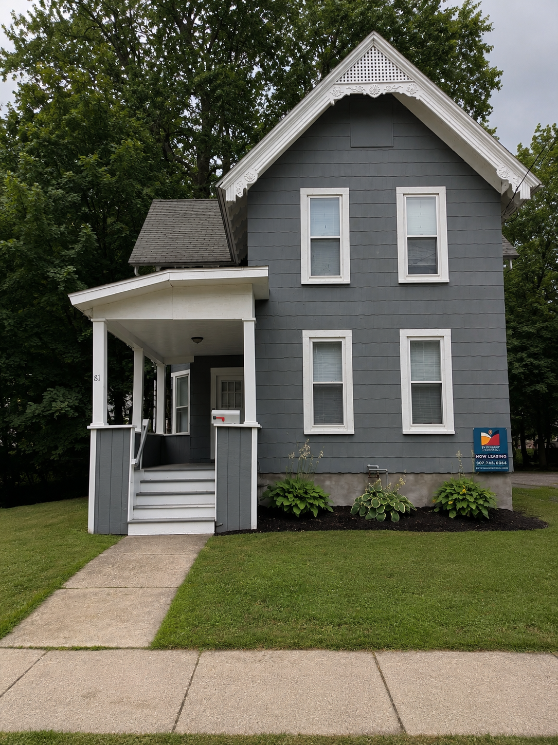 A three-story light gray house with a steep gabled roof, white trim, and a small covered front porch. The house has four windows visible on the front and a sign indicating student rentals.