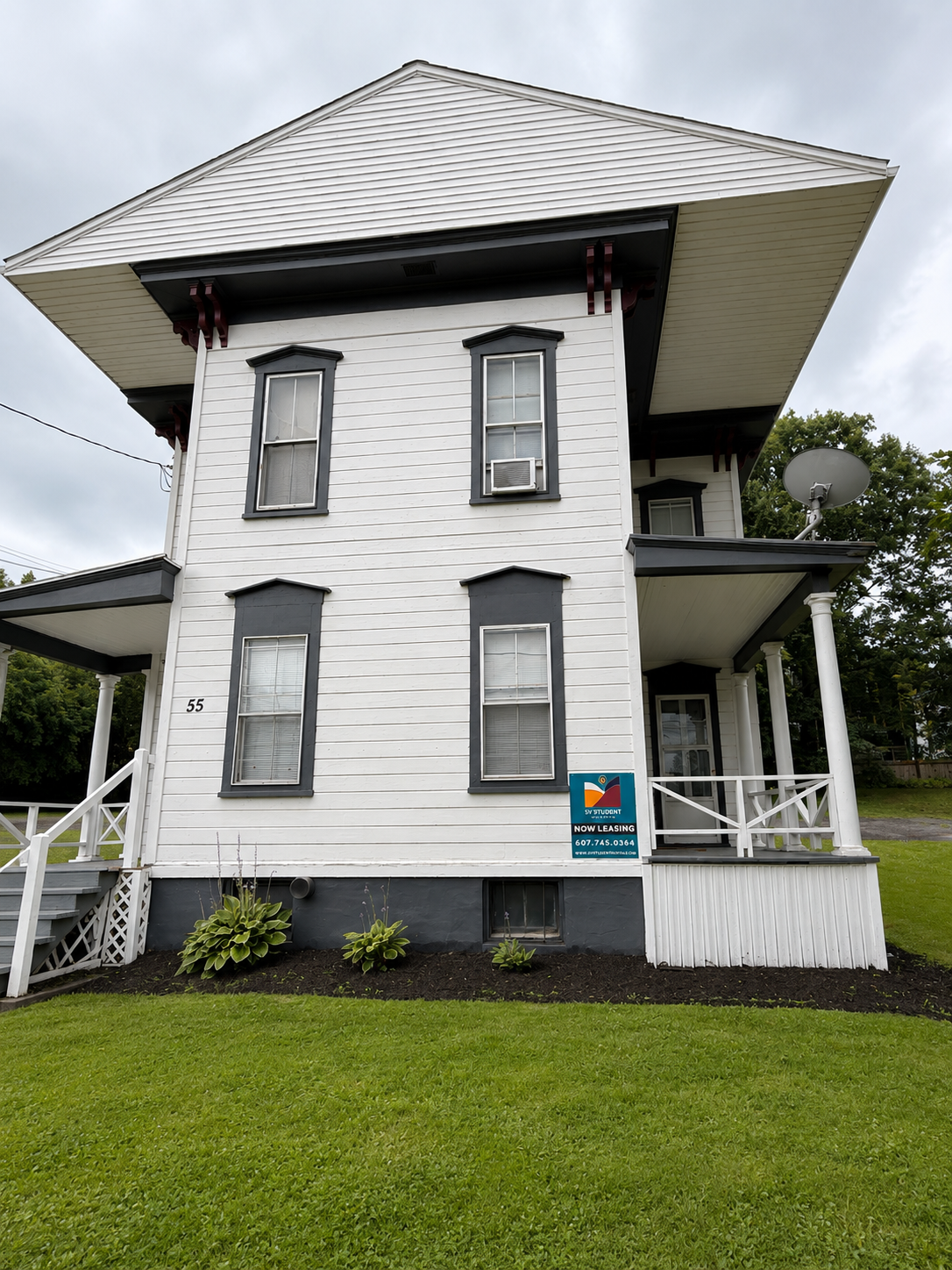 A three-story white house with black trim, a front staircase, and a small front yard. There are signs indicating the house is available for student rentals.