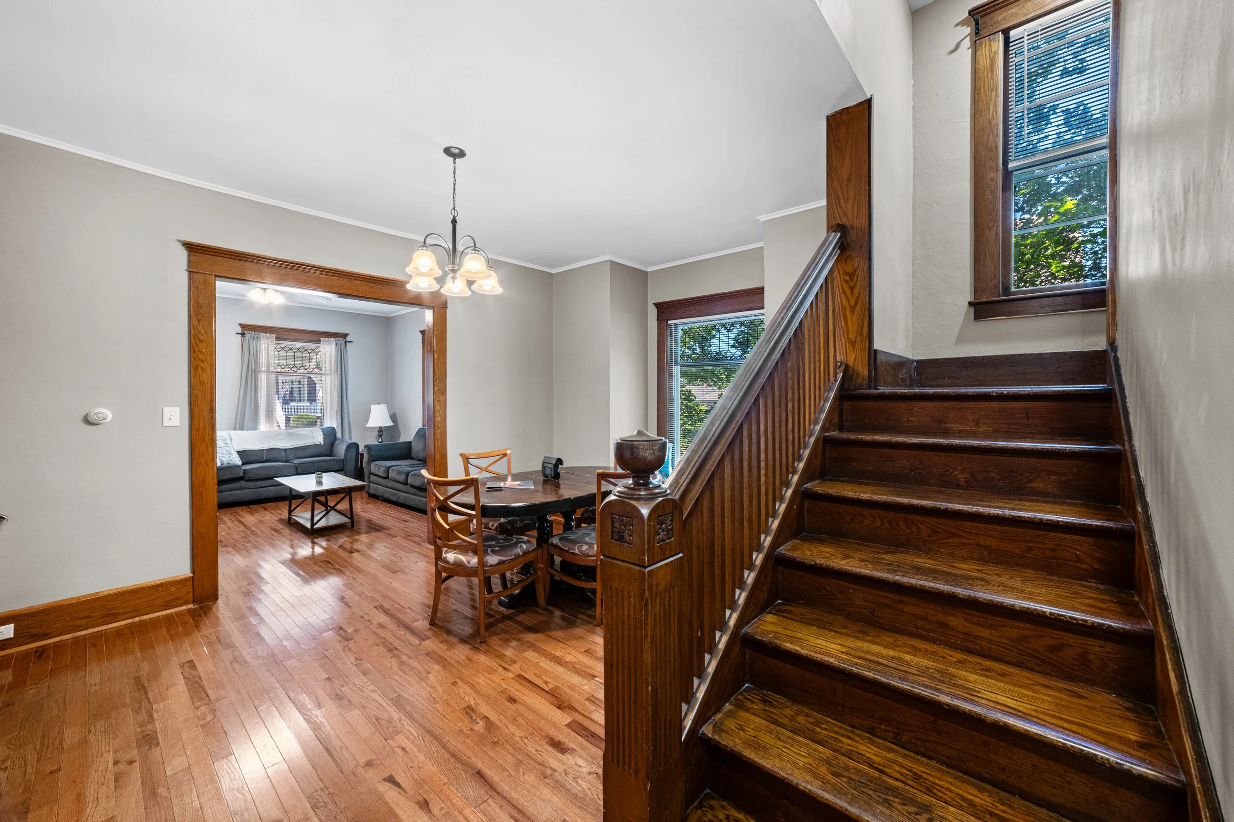 View of a living room and dining area with wooden floors, staircase, and windows with blinds, in a house interior.