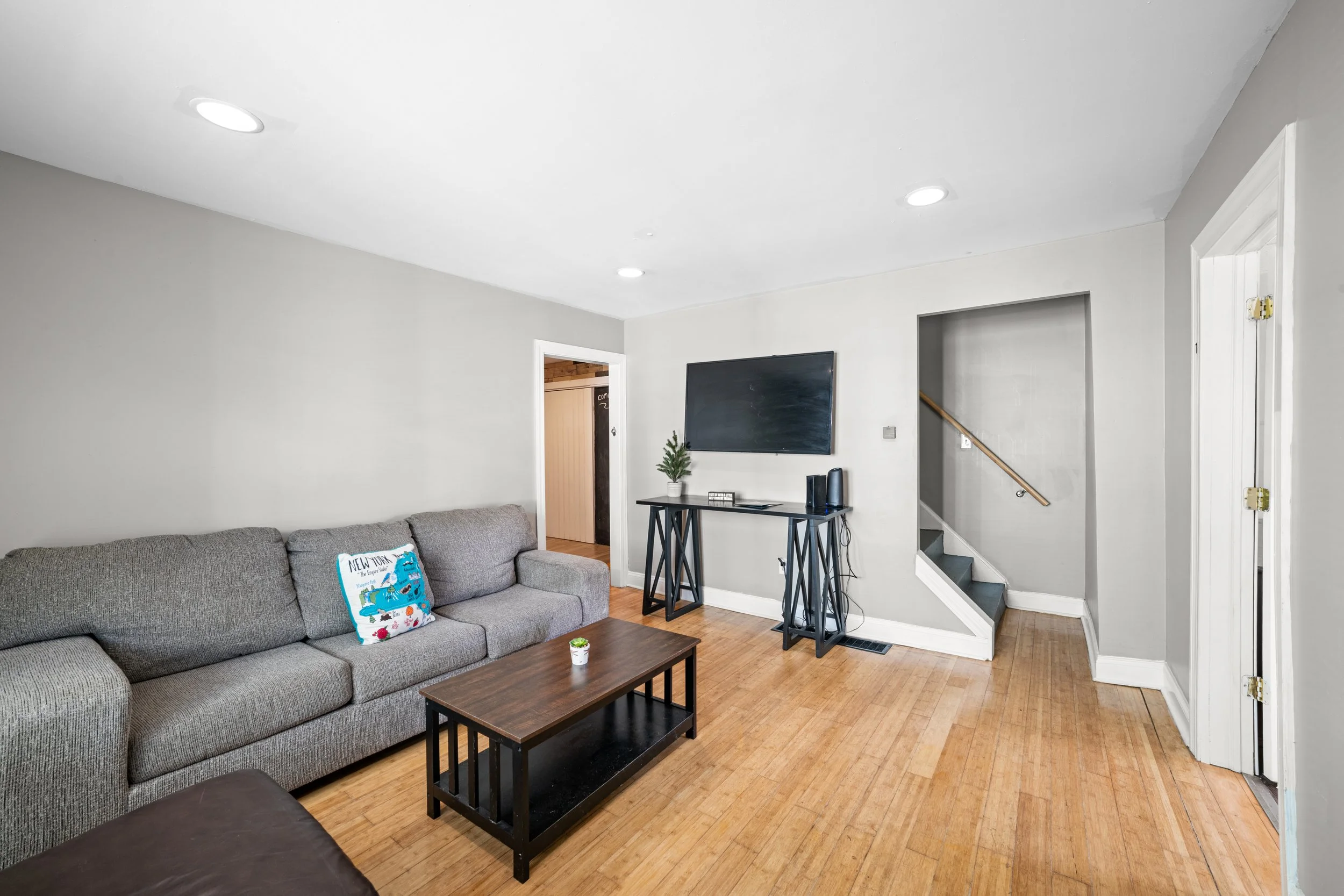 Living room with gray sofa, wooden floor, wall-mounted TV, small black table, and staircase leading upstairs.