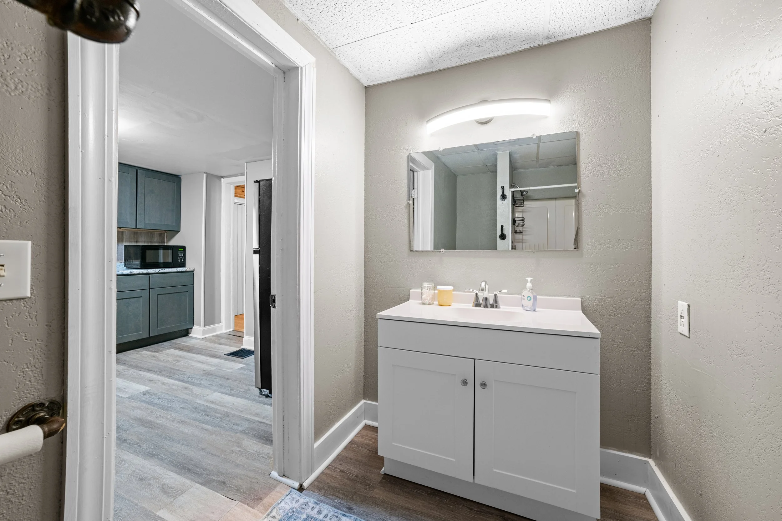 Bathroom with a white vanity, mirror, and soap dispenser, adjacent to a kitchen with grey cabinets and a microwave.