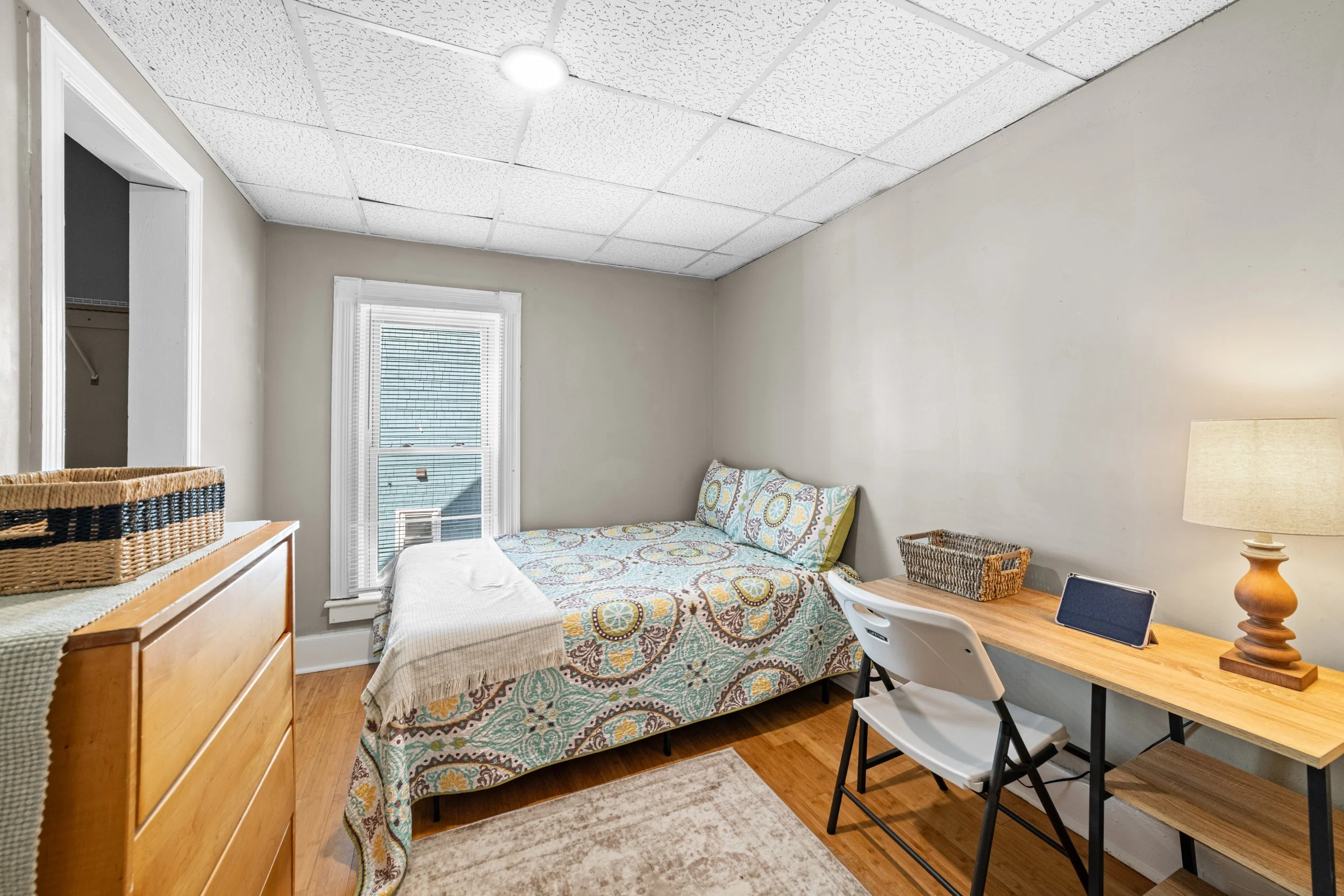 A bedroom with a bed, dresser, desk with a lamp, and a window, featuring beige walls and hardwood floors.