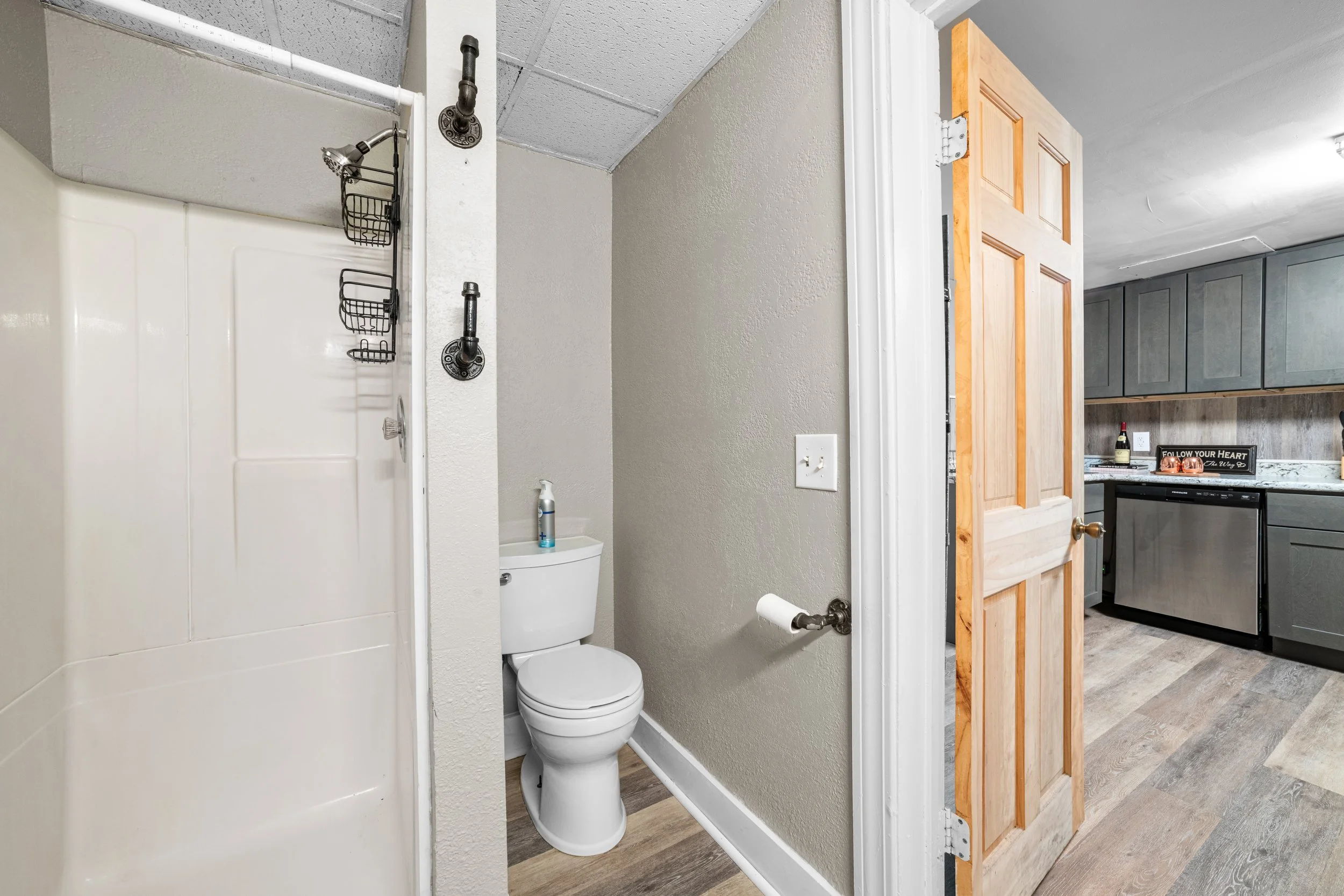 Small bathroom with a toilet and shower, visible from an open door leading to a kitchen area.