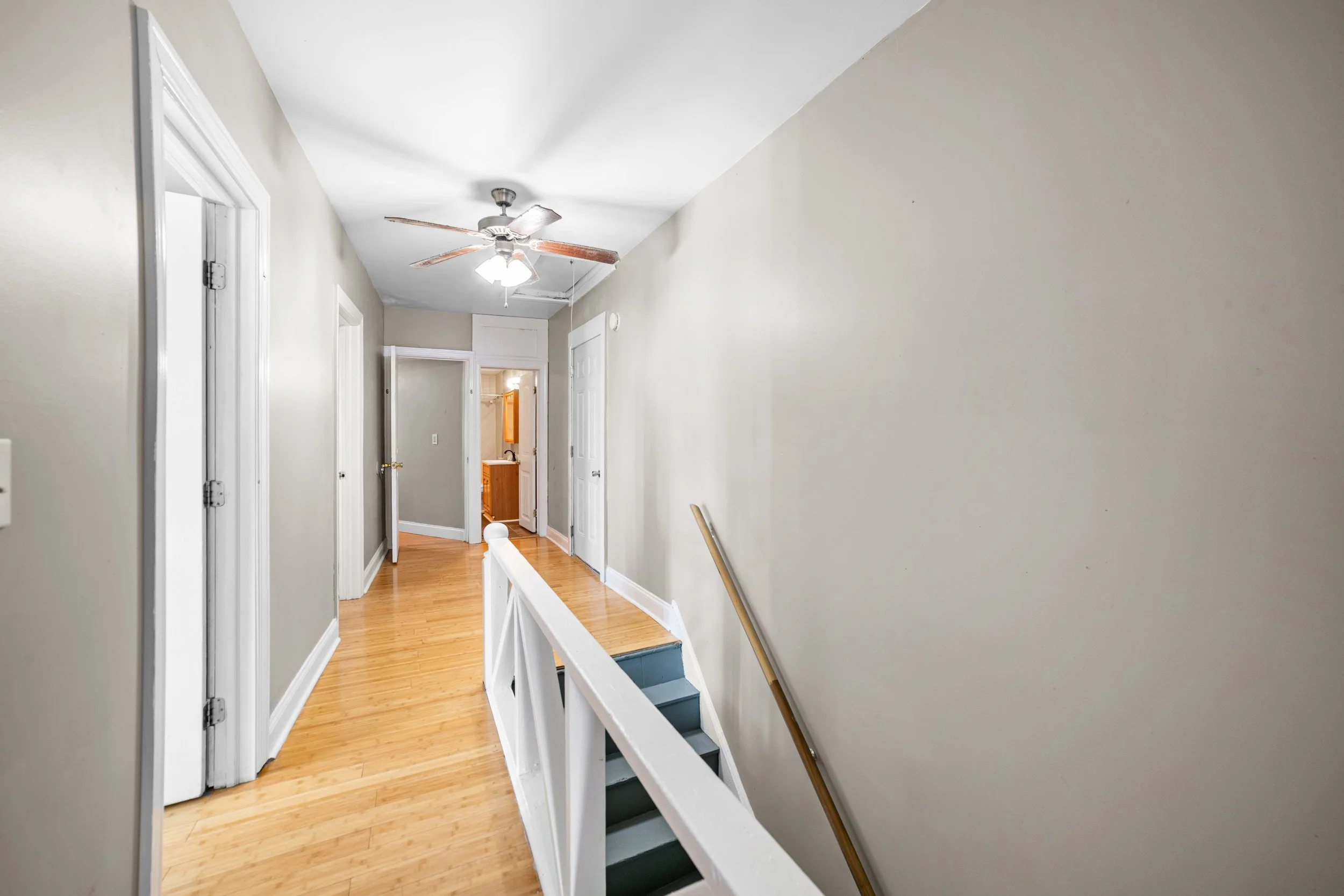 Upward view of a hallway with wooden flooring, white walls, multiple closed doors, a ceiling fan with lights, and a staircase with a wooden handrail.