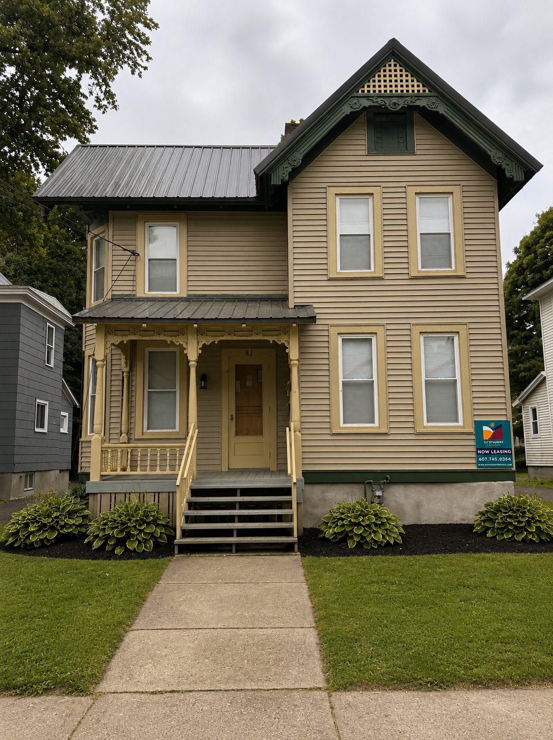 Green three-story Victorian house with a metal roof, a front porch with decorative trim, and several windows framed in beige, surrounded by trees and neighboring houses.