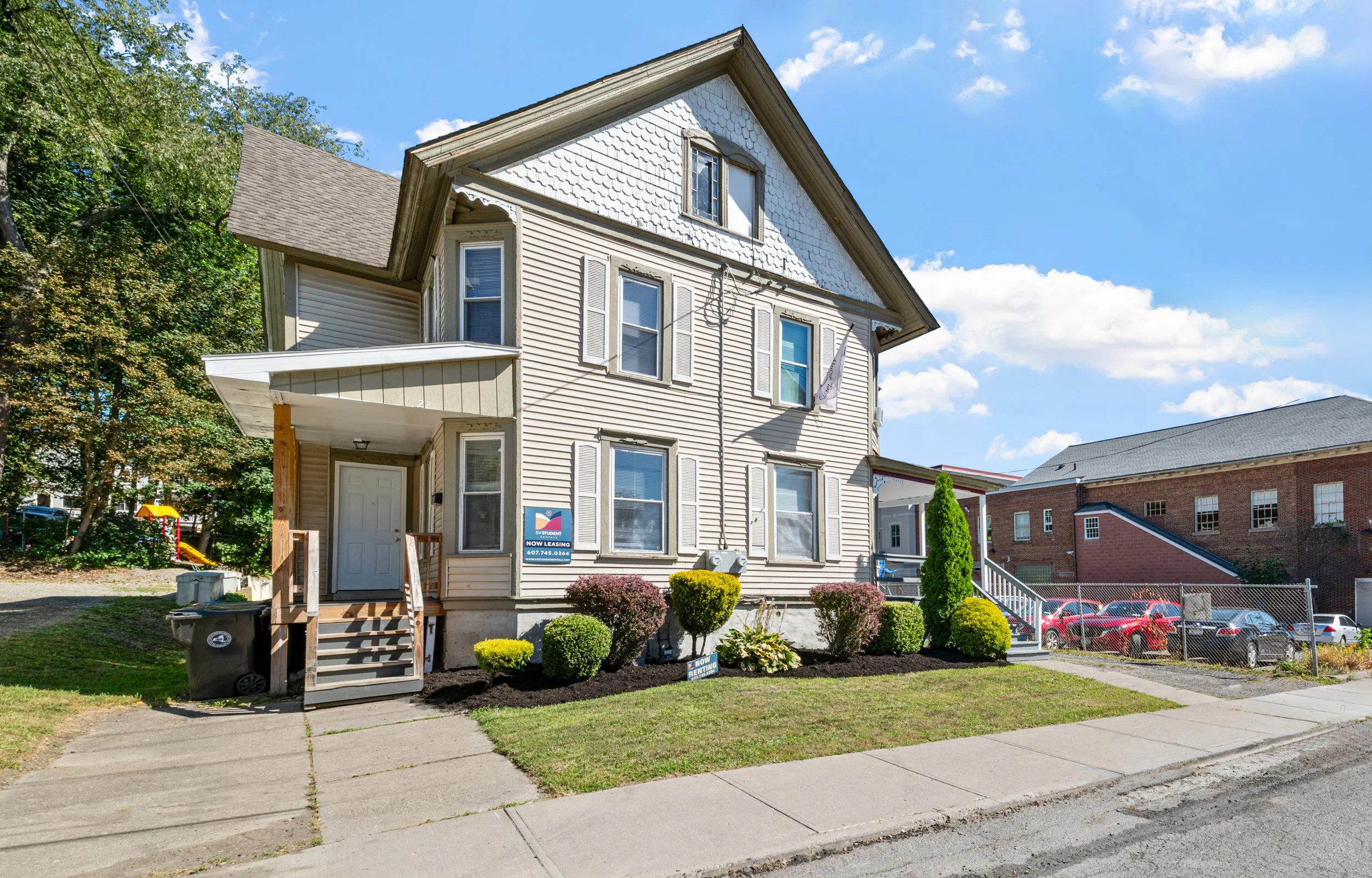 Multi-story beige house with a front porch, surrounded by a well-manicured lawn and small bushes, with a blue sky and some cars in the background.