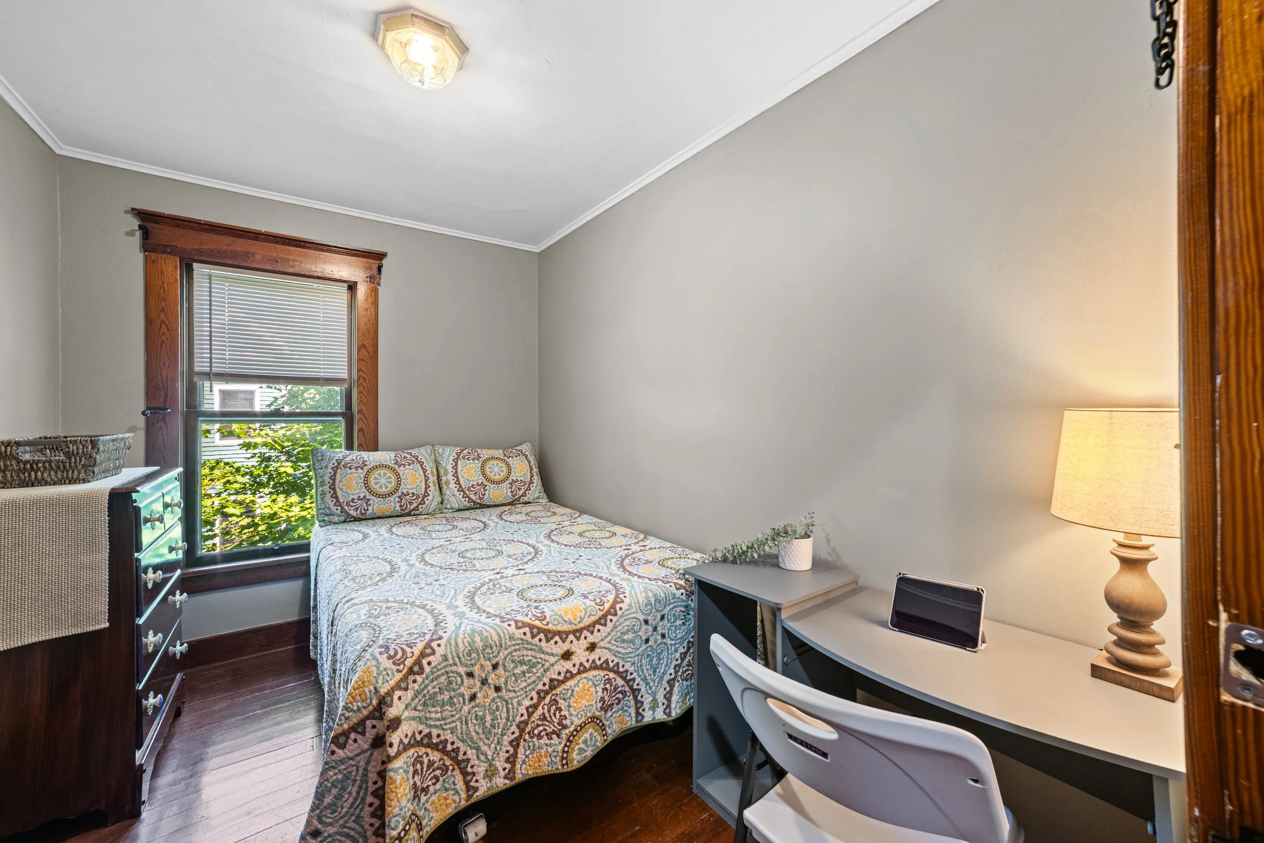 Small bedroom with a bed, desk, and dresser, window with blinds, wooden trim, and a lamp on the desk.