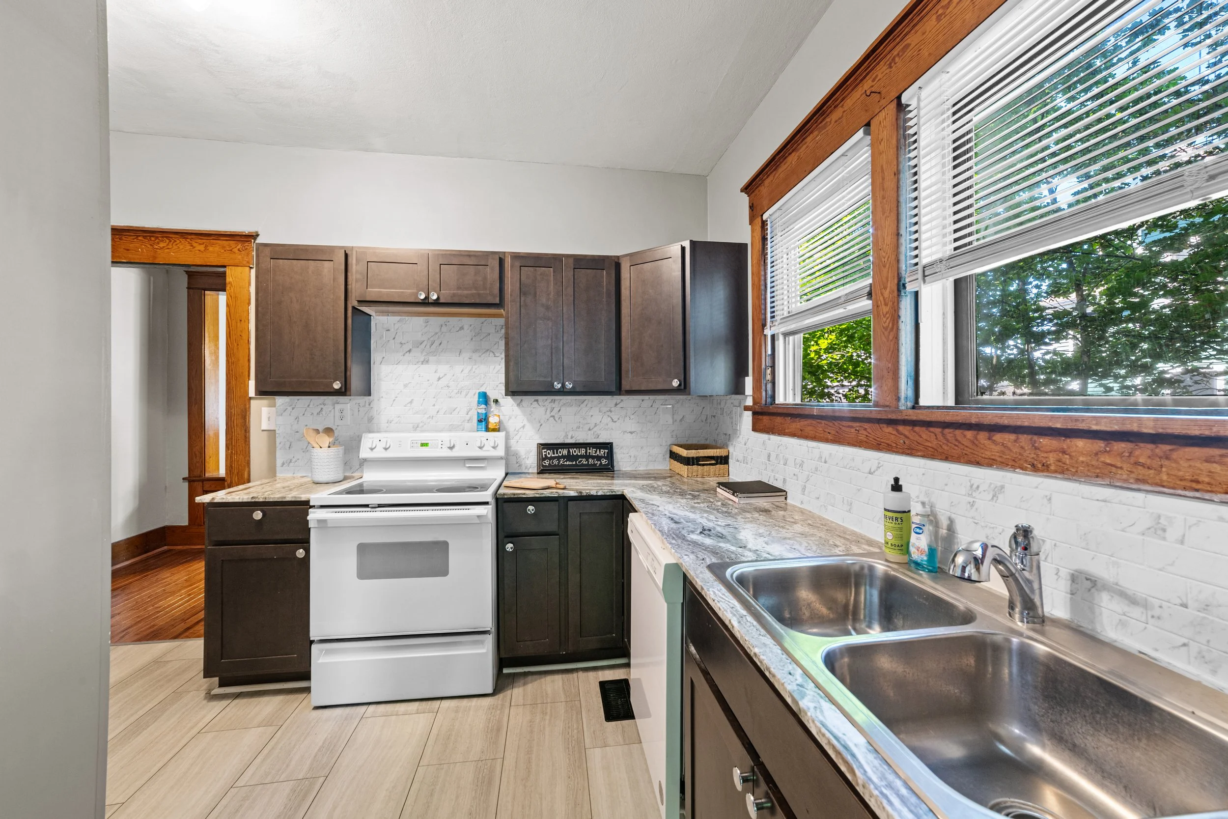 Kitchen with white stove, brown cabinets, granite countertops, double sink, large window with blinds, and a small sign that reads 'Follow Your Heart'.