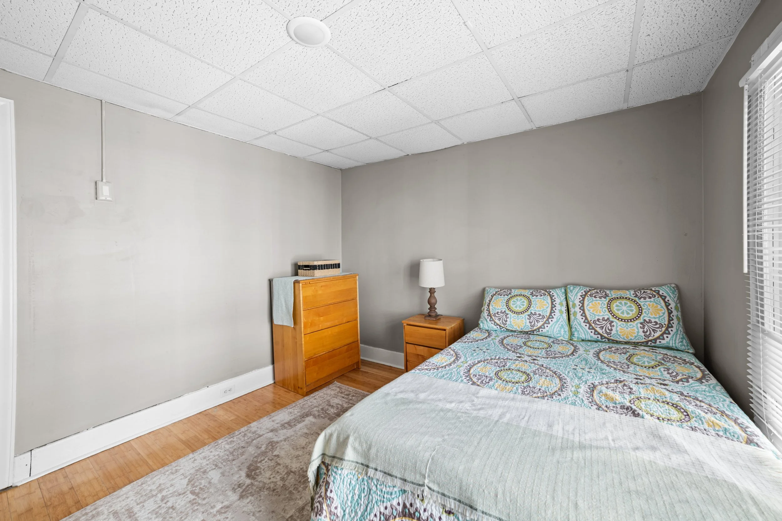 A simple bedroom with a bed, a nightstand with a lamp, and a dresser against a gray wall. There is a window with blinds on the right and a beige carpet on hardwood floors.