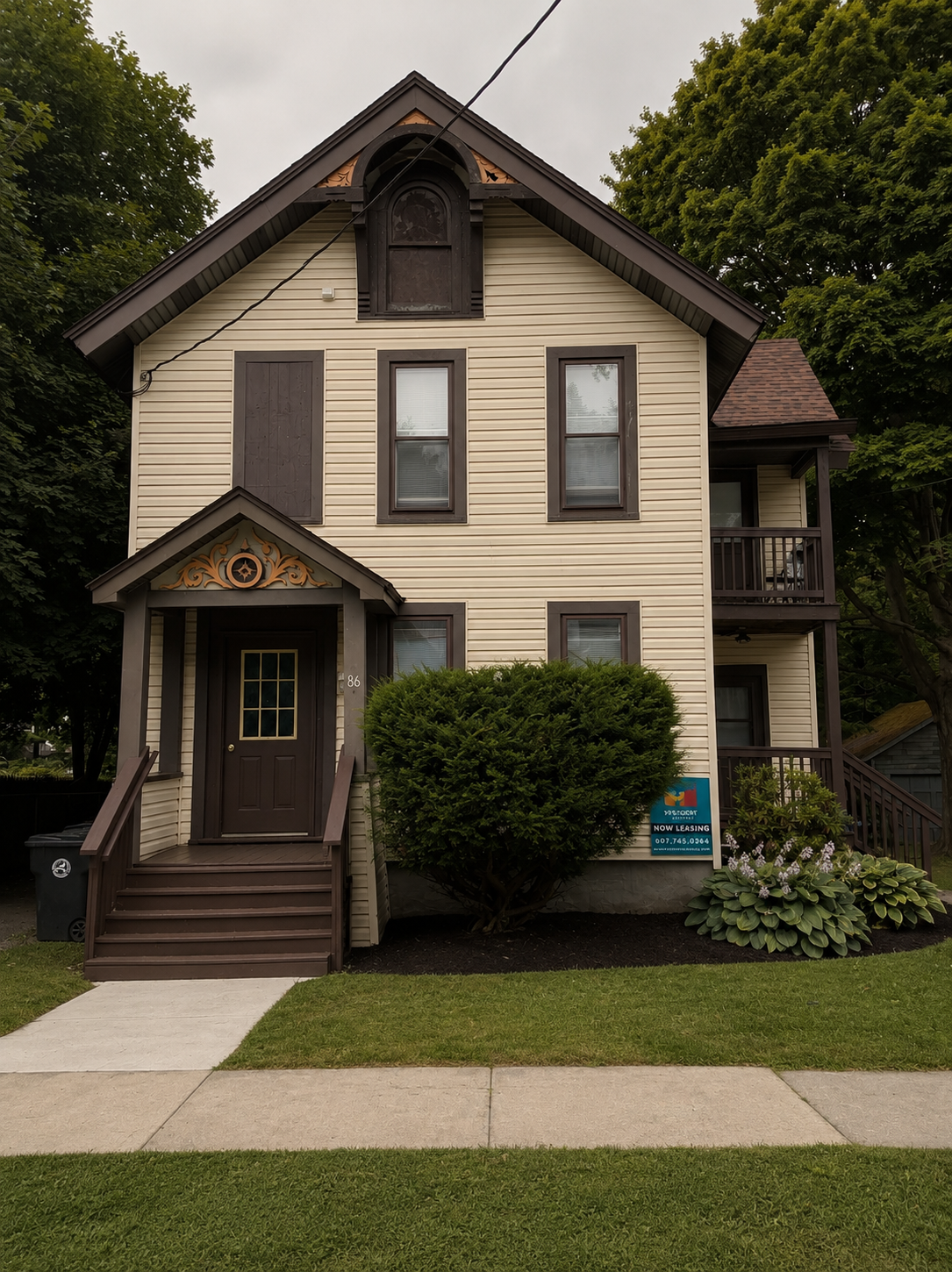 Three-story house with beige siding, dark brown trim, front steps, and a small porch. The house has tall windows with blinds, a small balcony on the right side, and a landscaped front yard with shrubs and grass. It appears to be undergoing some repairs or renovation, as there is a boarded-up window and decorative woodwork around the entrance and attic.