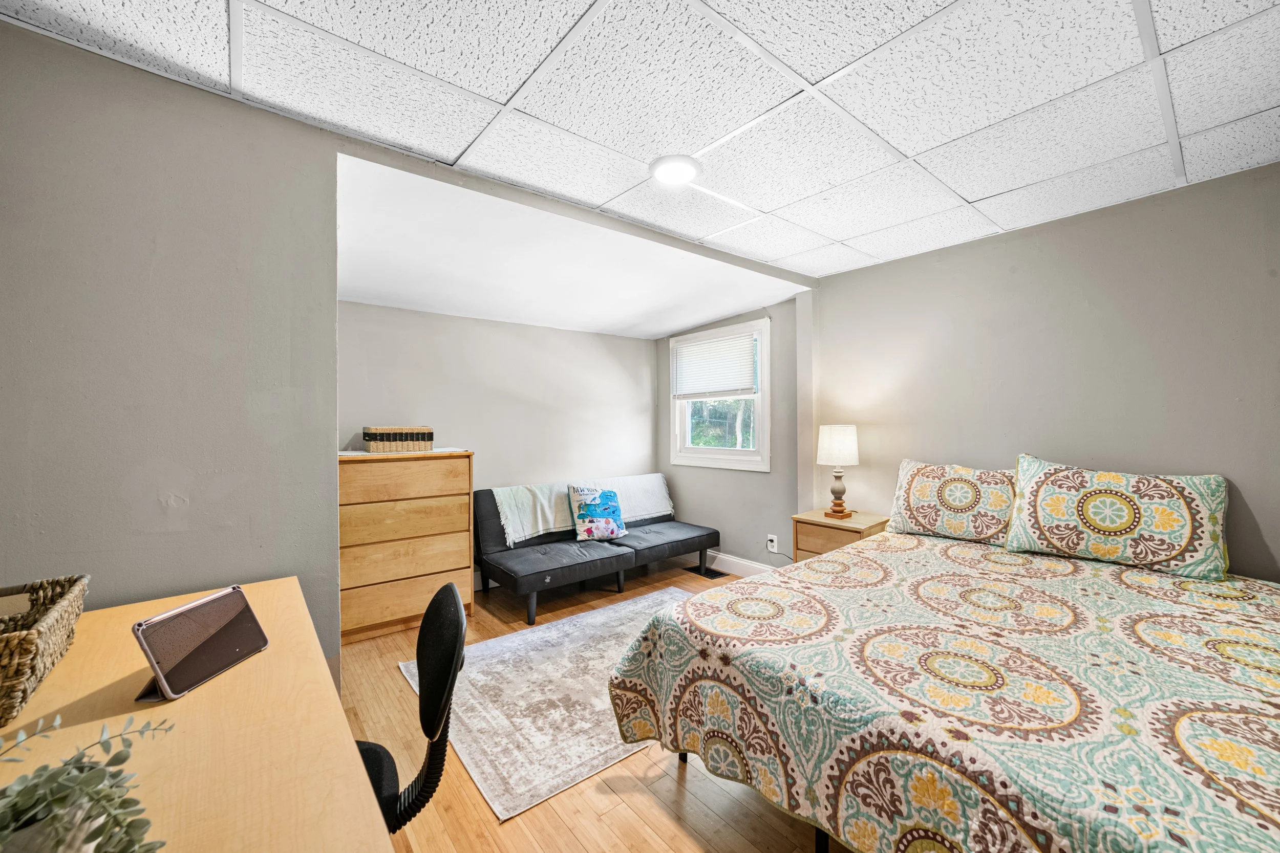 A bedroom with a bed covered in a multicolored patterned quilt, a nightstand with a lamp, a black futon with pillows, a dresser, a wooden table, a chair, and a window letting in natural light.