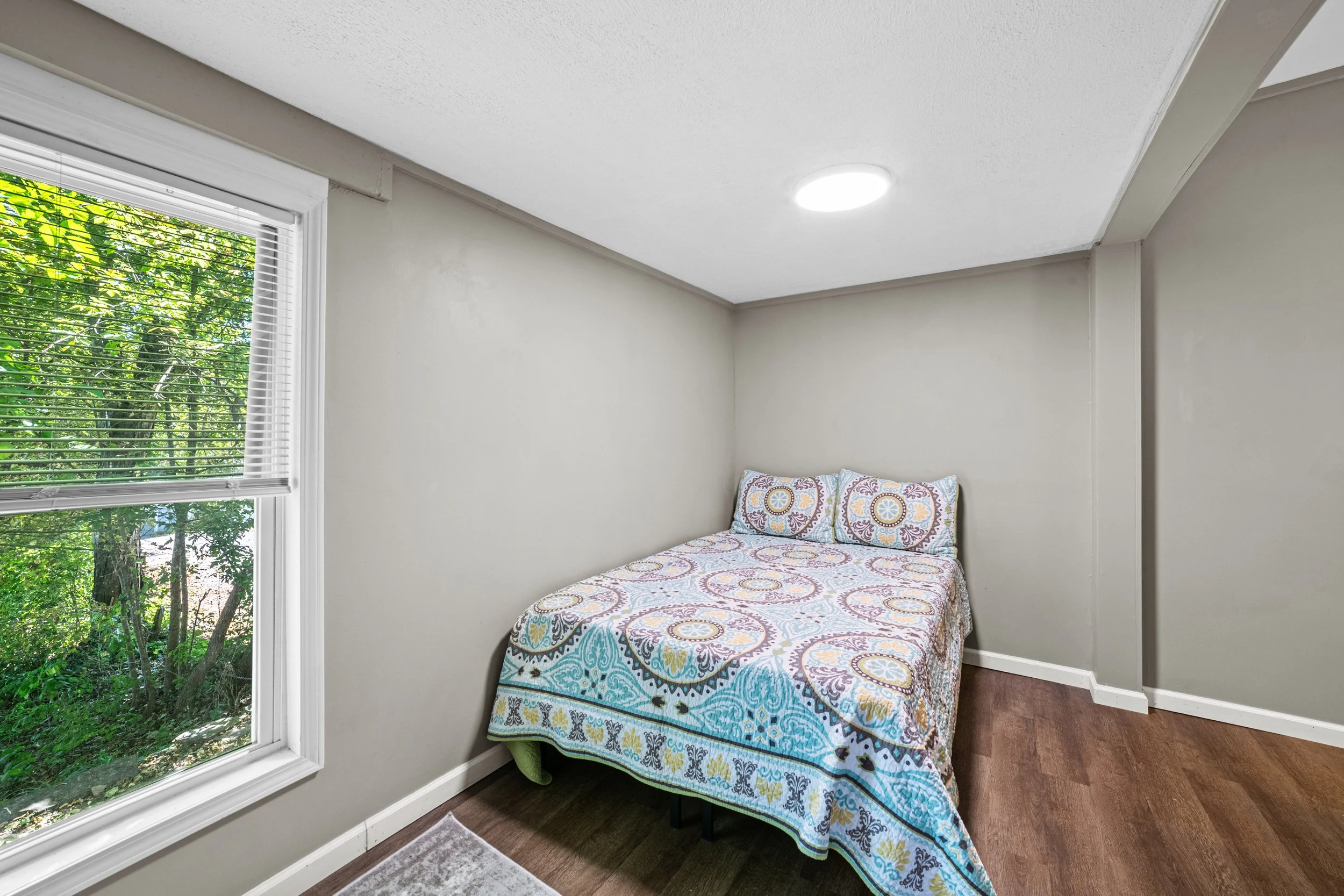 Small bedroom with a window, a bed with colorful patterned bedding and pillows, and hardwood flooring.