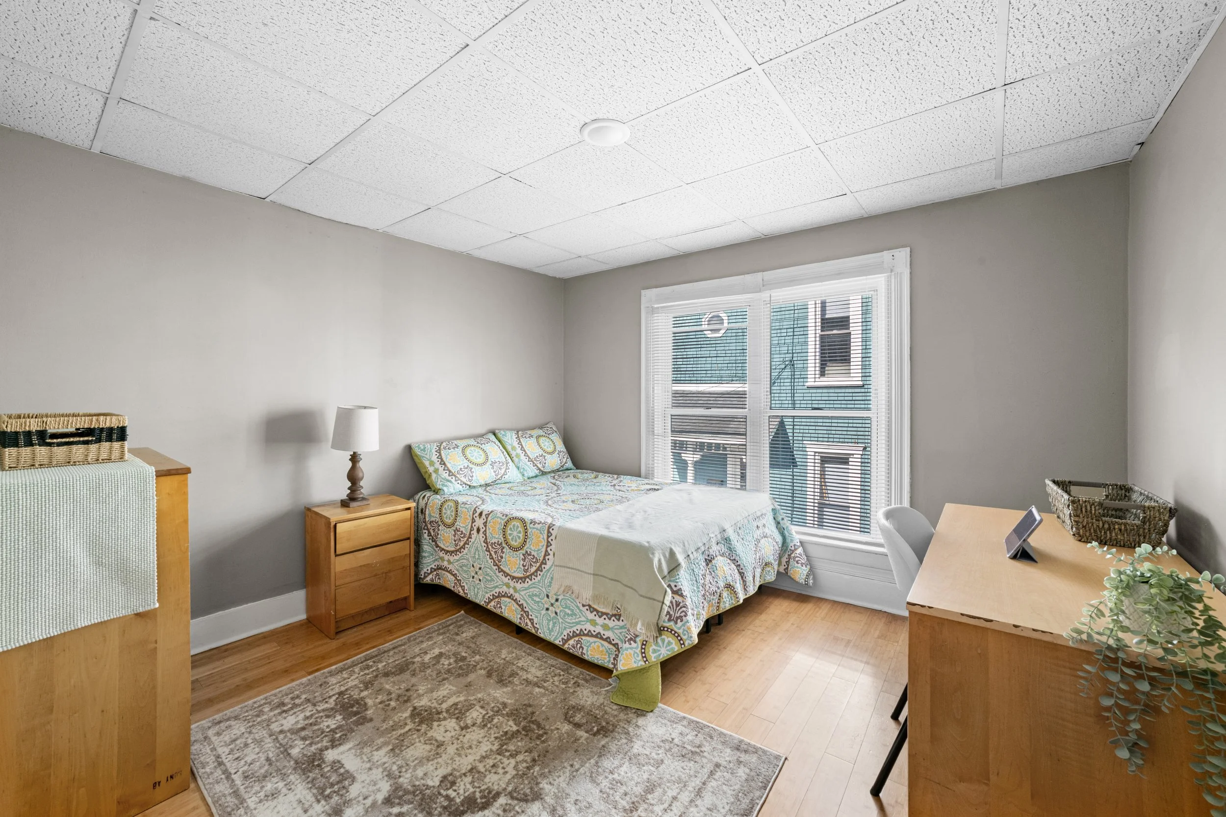 A small bedroom with a bed, nightstand, and dresser. The room has hardwood floors, a large window with blinds, and a textured ceiling.