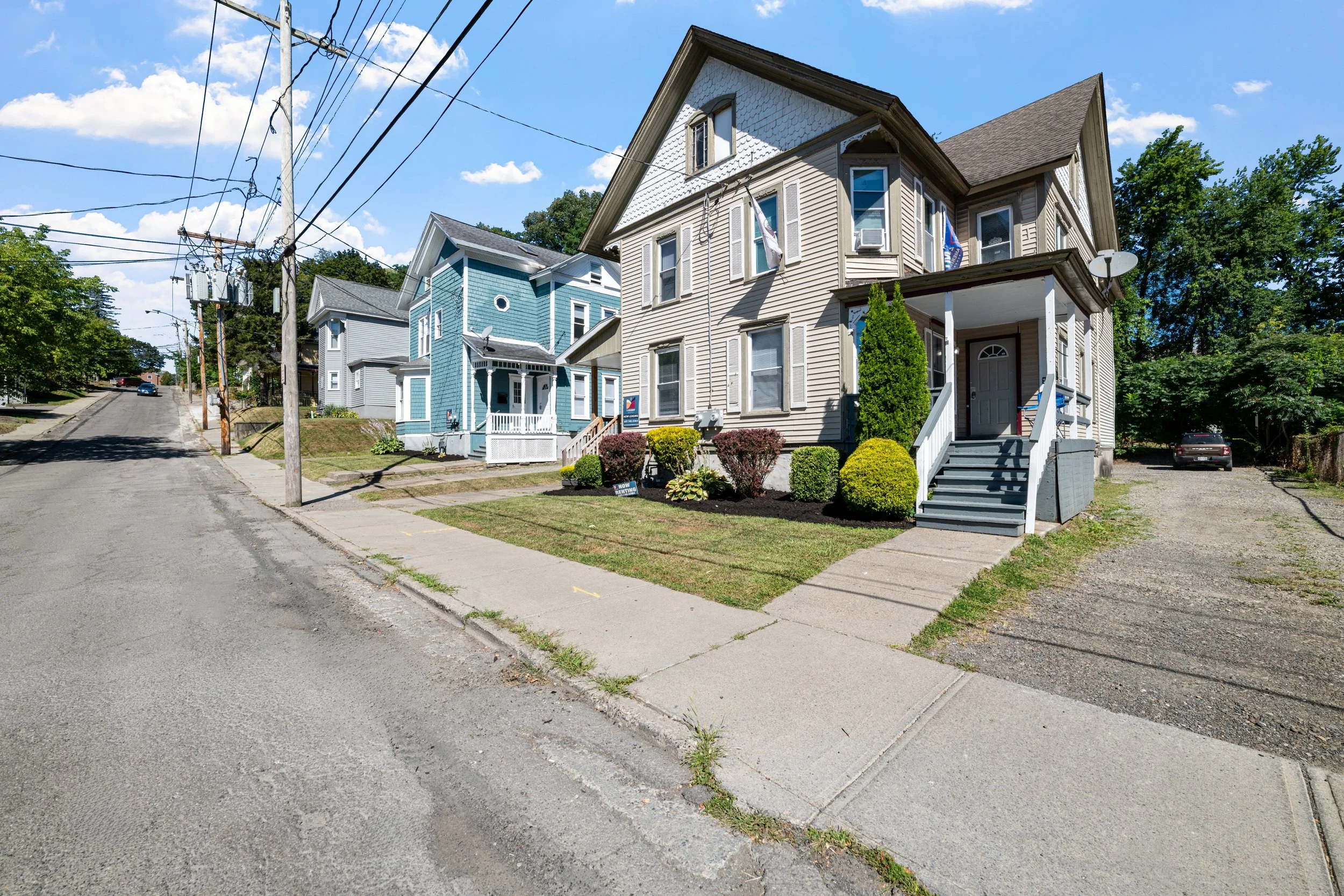 Residential street with multiple colorful houses, including a beige house with stairs to the front door, greenery, and an overcast sky.