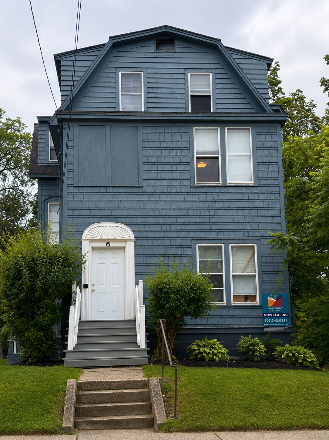 A three-story blue house with white stairs leading to a front door, multiple windows, and a sign indicating it is available for student rentals.