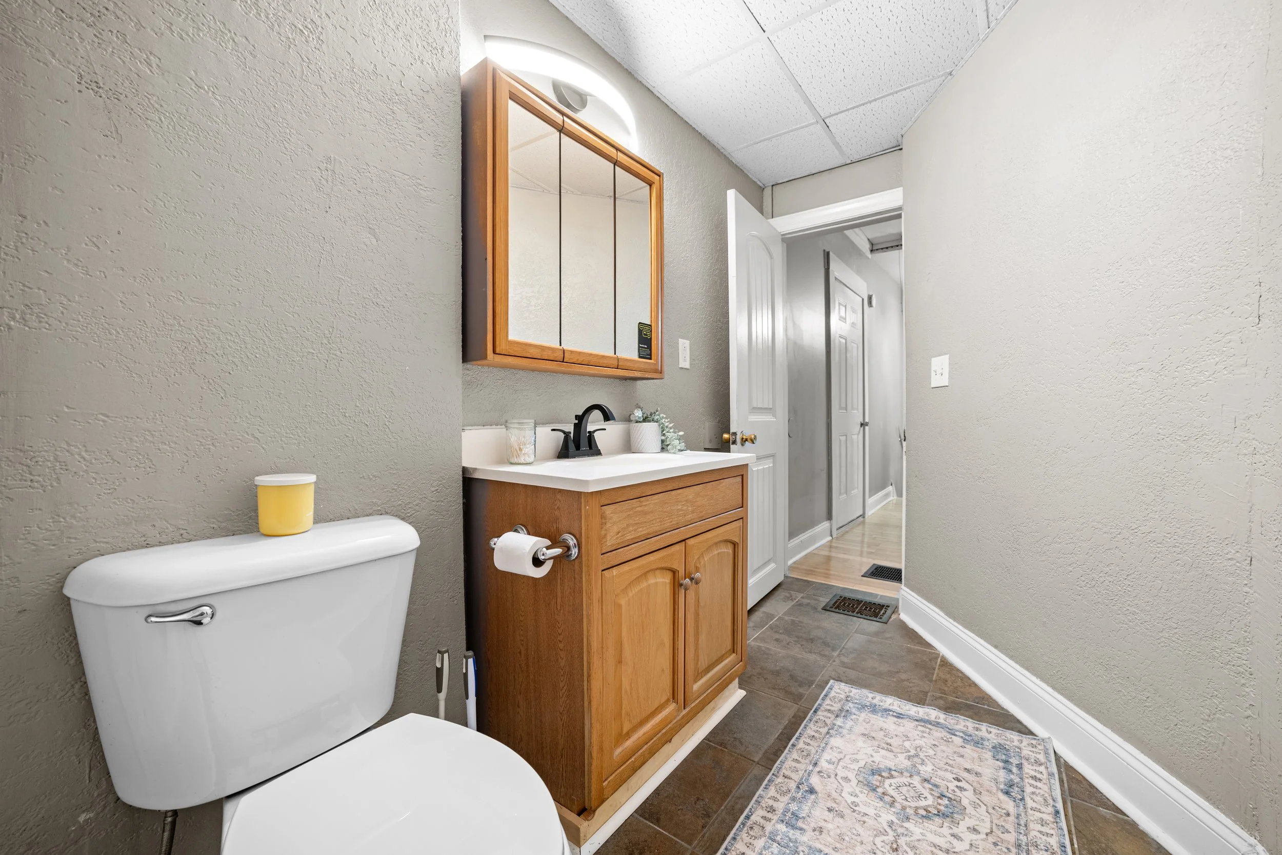 Bathroom with a toilet, wooden vanity with sink, mirror cabinet, and a small rug on brown tile floor, hallway visible in the background.