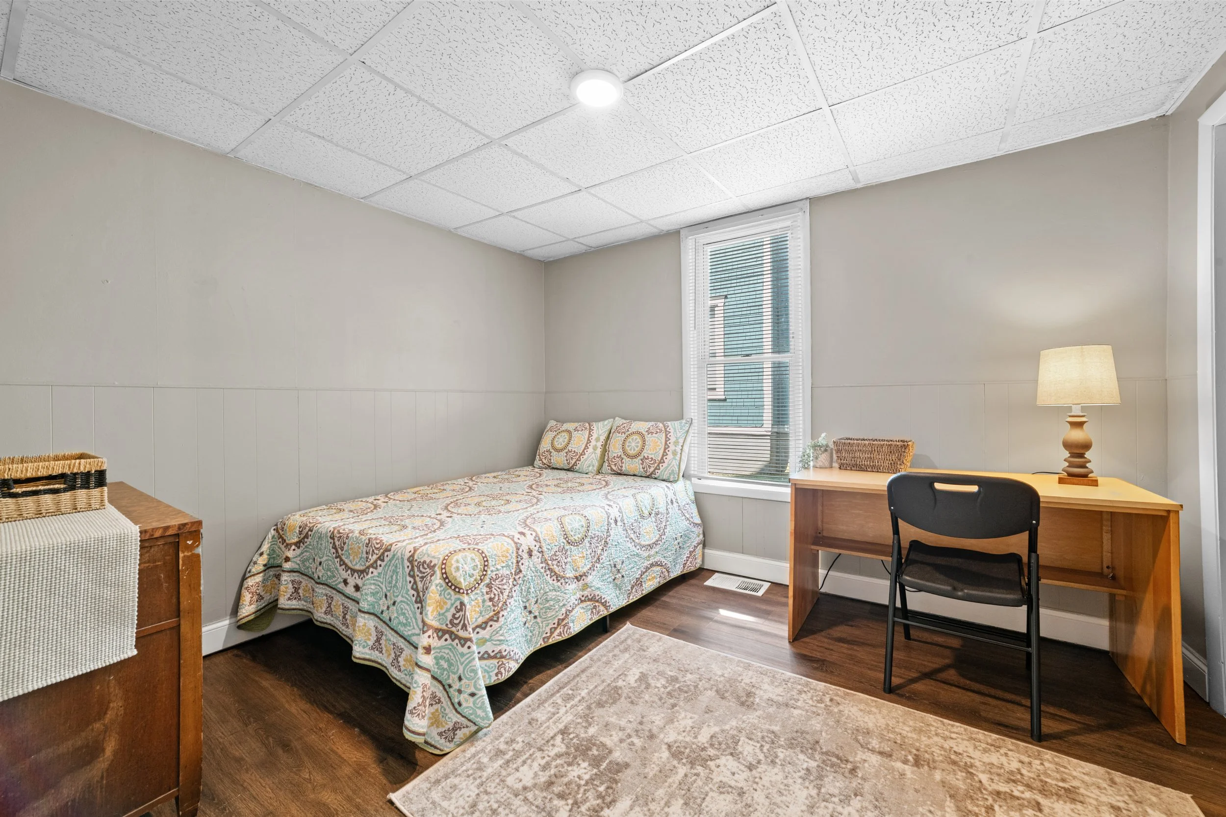 A simple bedroom with a bed, table, and chair, featuring a window with blinds, a lamp, and a decorative rug on the wooden floor.