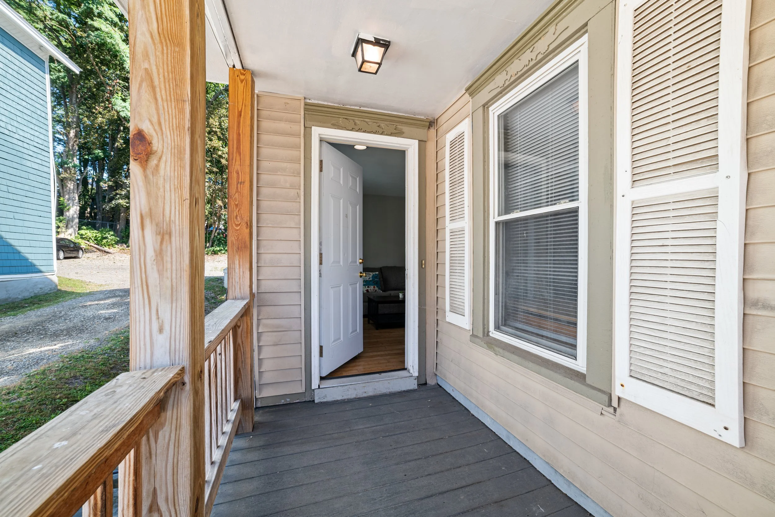 View of a front porch with wooden railing, leading to an open door showing a room with a gray couch and wooden floor inside. The house's beige exterior has two white-framed windows with white shutters, and a ceiling light fixture.