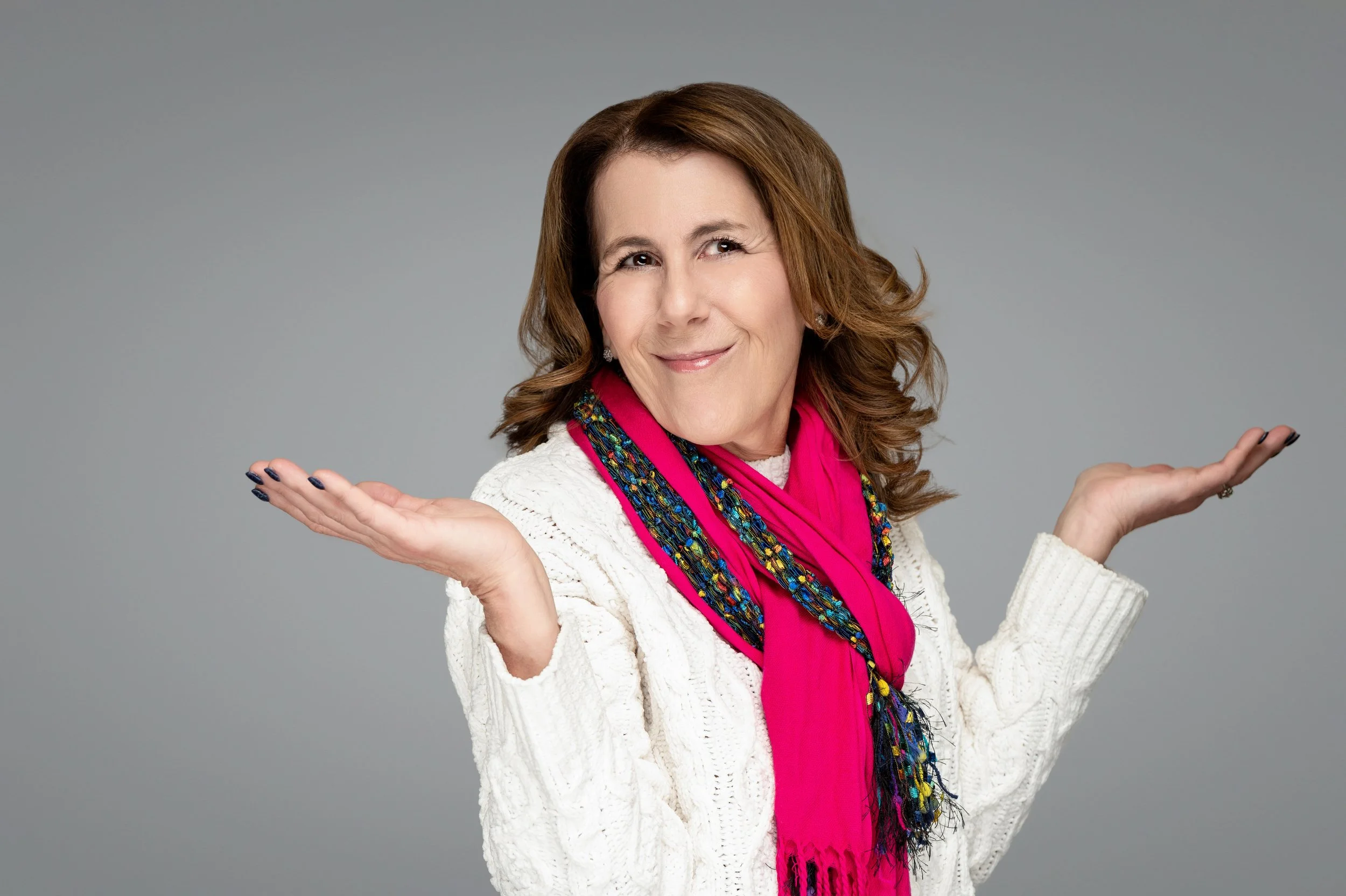 Beth Blacker smiling with both hands raised in an open, welcoming gesture, wearing a white sweater and bright pink scarf against a neutral gray background.