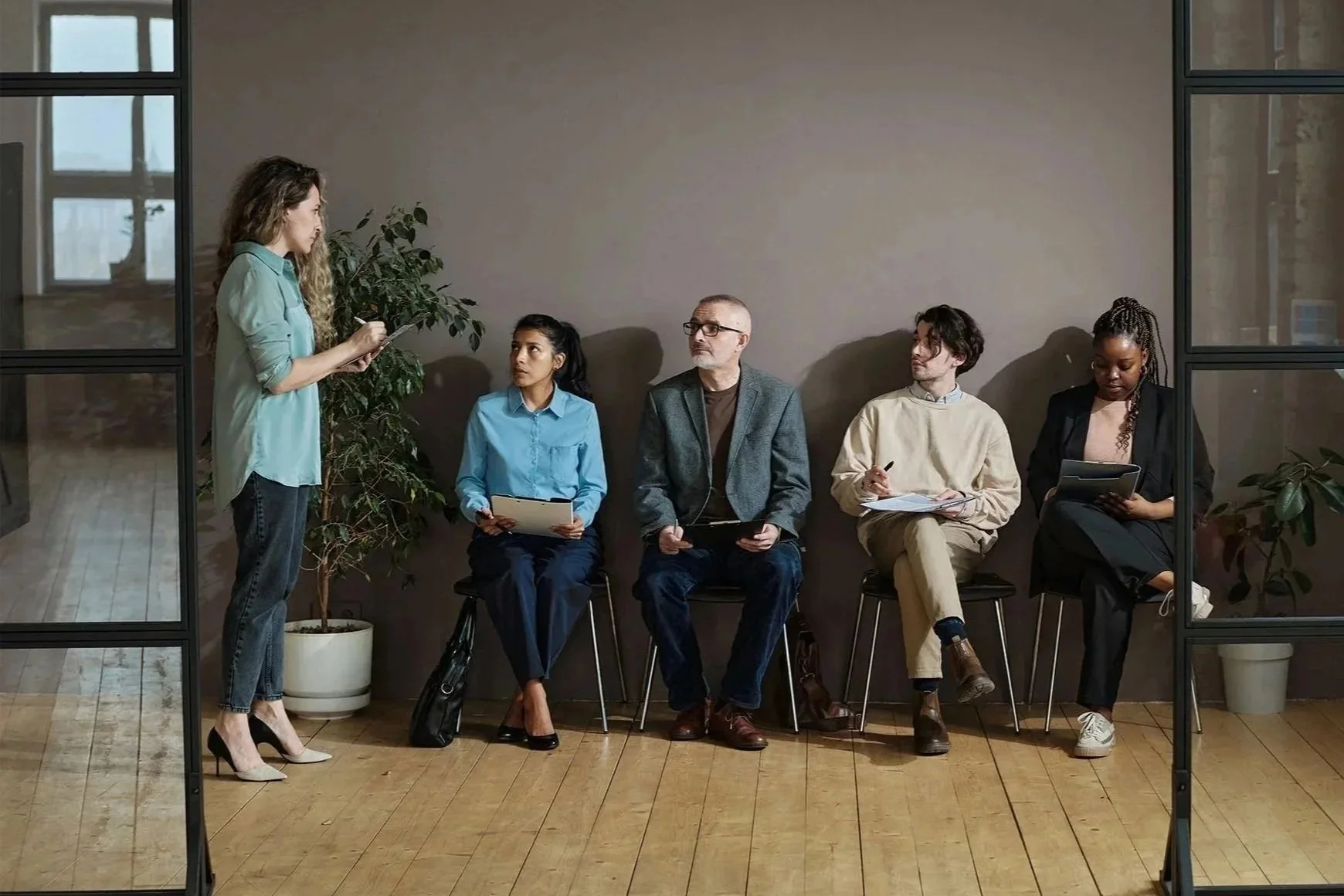 A woman standing and speaking to four seated people in a modern office or conference room with large glass panels. The seated individuals are taking notes or listening attentively.