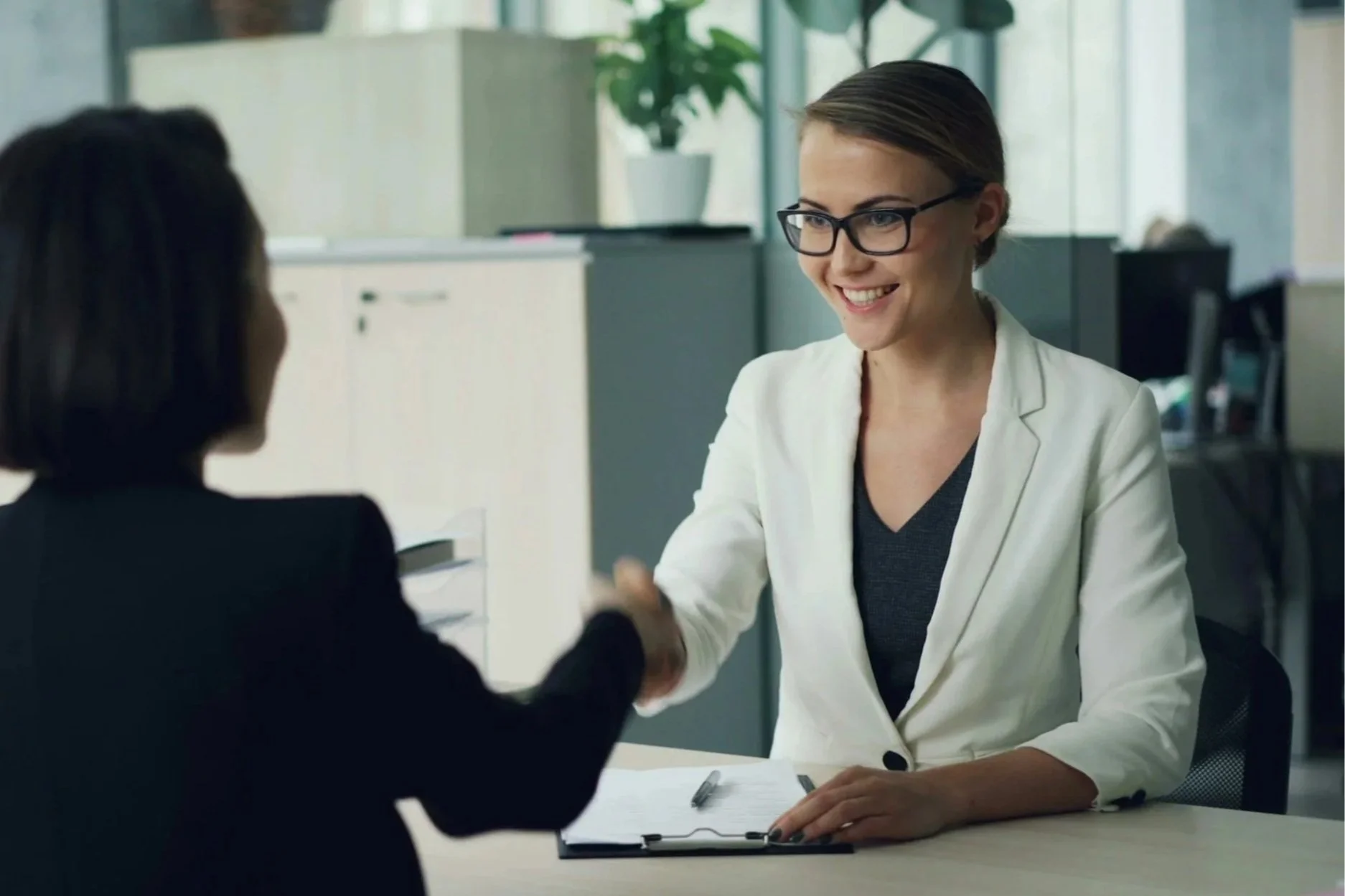 Two women in a professional setting shaking hands across a table, one wearing glasses and a white blazer, the other in a dark suit, with a clipboard and pen on the table.