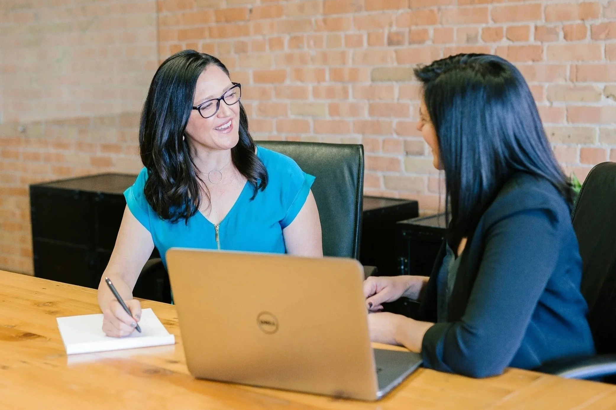 Two women sitting at a wooden table, engaged in a conversation, with a laptop and notepad between them in a modern office with exposed brick walls.