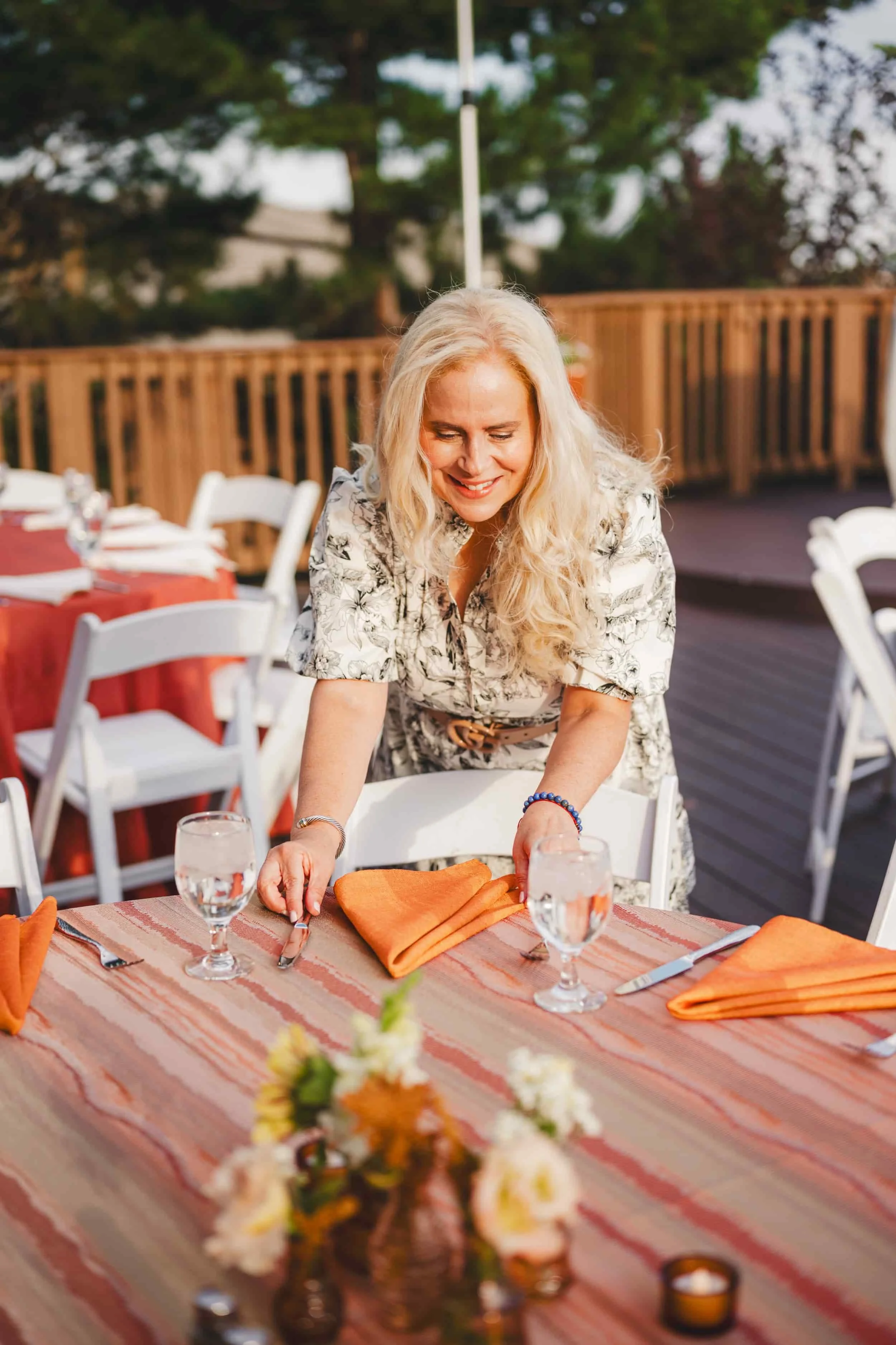 A woman with blonde hair smiling and setting a table outdoors with orange napkins, wine glasses, and floral centerpieces created by exceptional event coordinator Kristen Collins with Typecast Meetings and Events