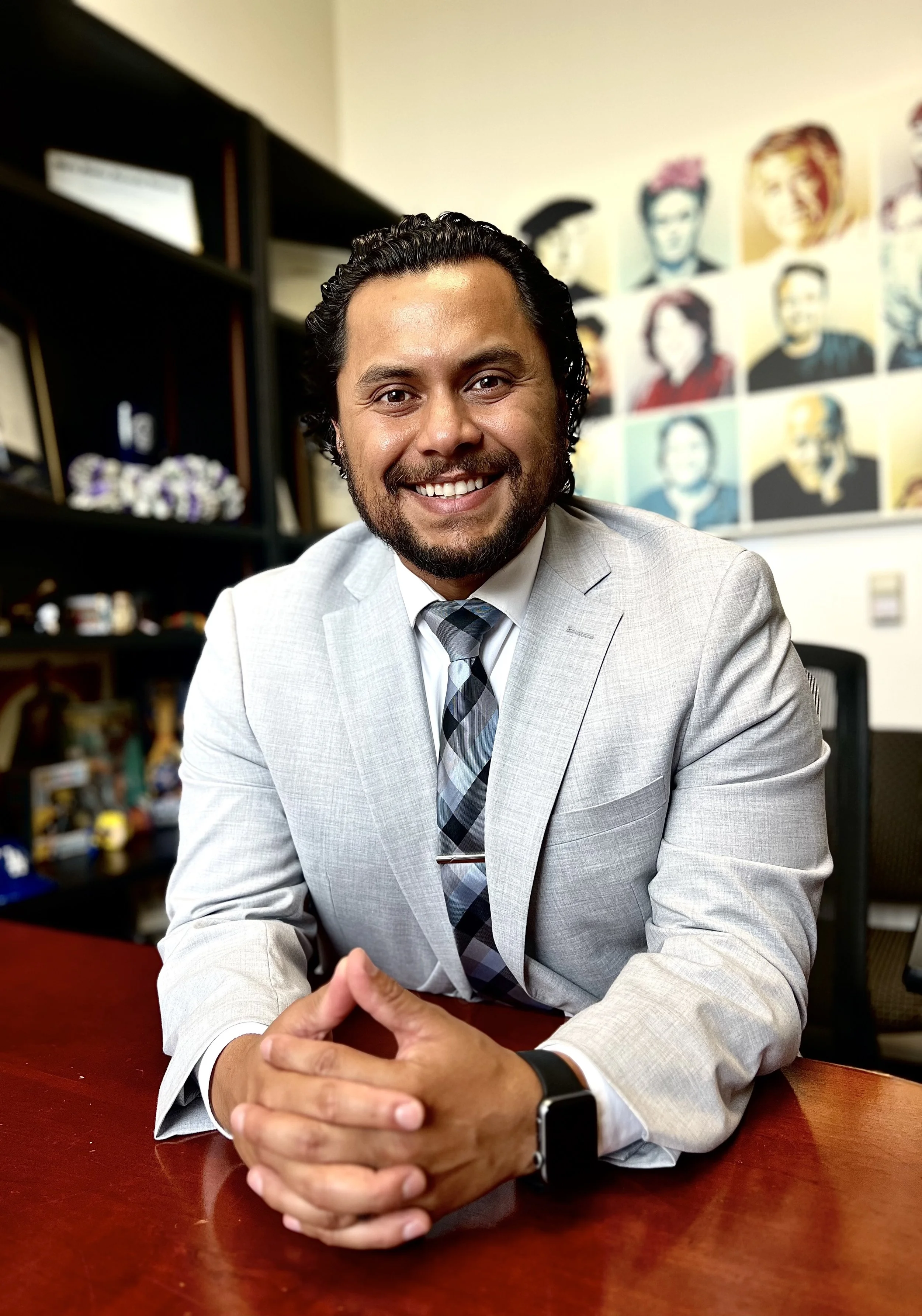 A man in a light gray suit and patterned tie sitting at a dark wooden desk with hands clasped. He is smiling and looking at the camera. In the background, colorful portraits are visible on the wall.