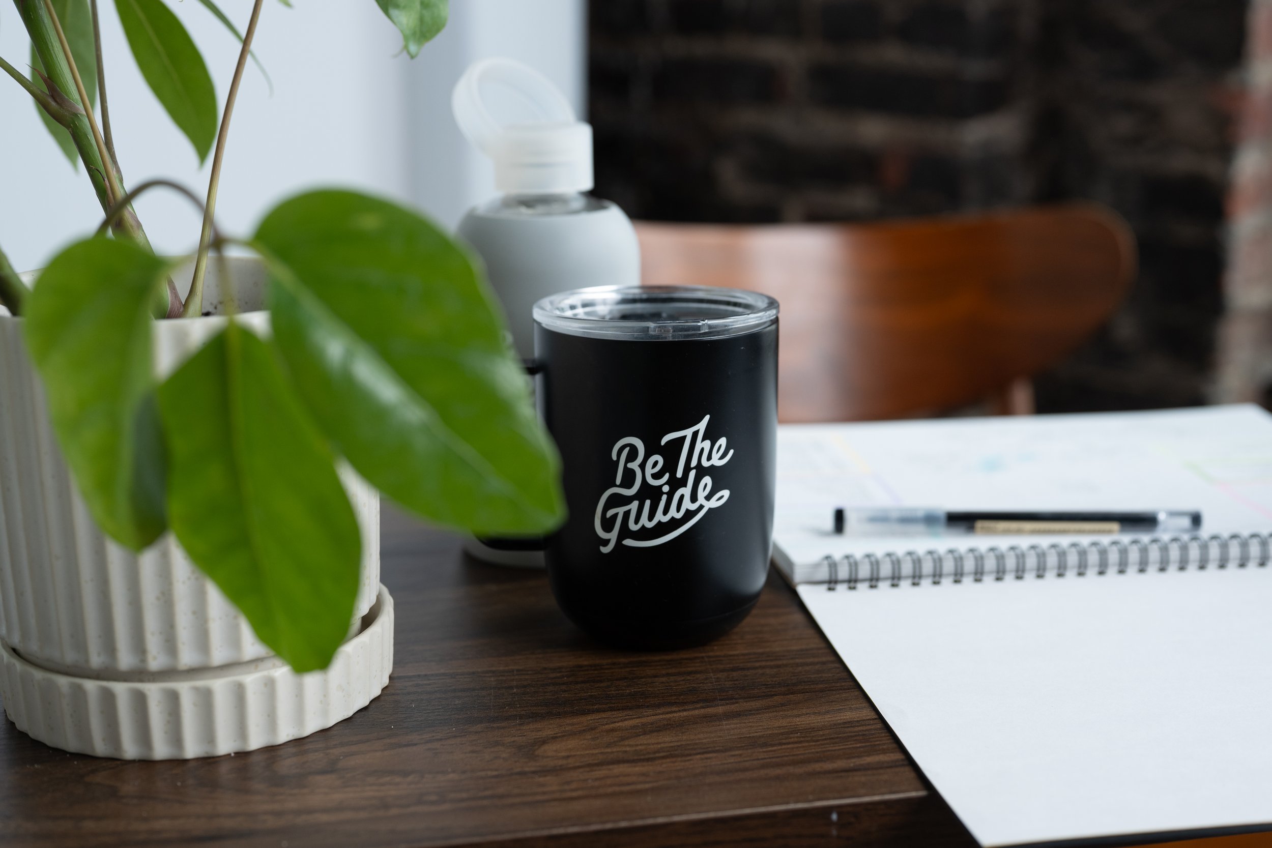 Desk scene with a notebook, pen, plant, and a mug that reads ‘Be the Guide,’ representing a customer-centric approach to content