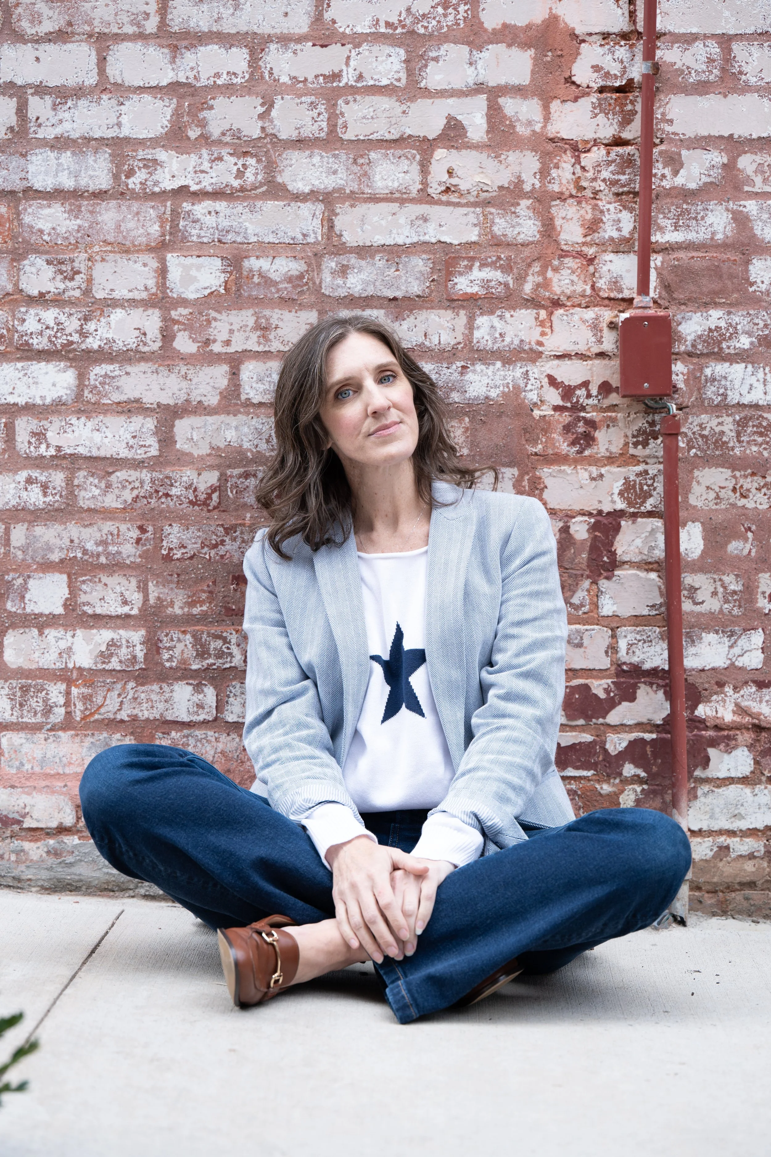 Annie Figenshu sitting cross-legged in front of a brick wall, wearing a light blazer and jeans, looking calmly toward the camera.