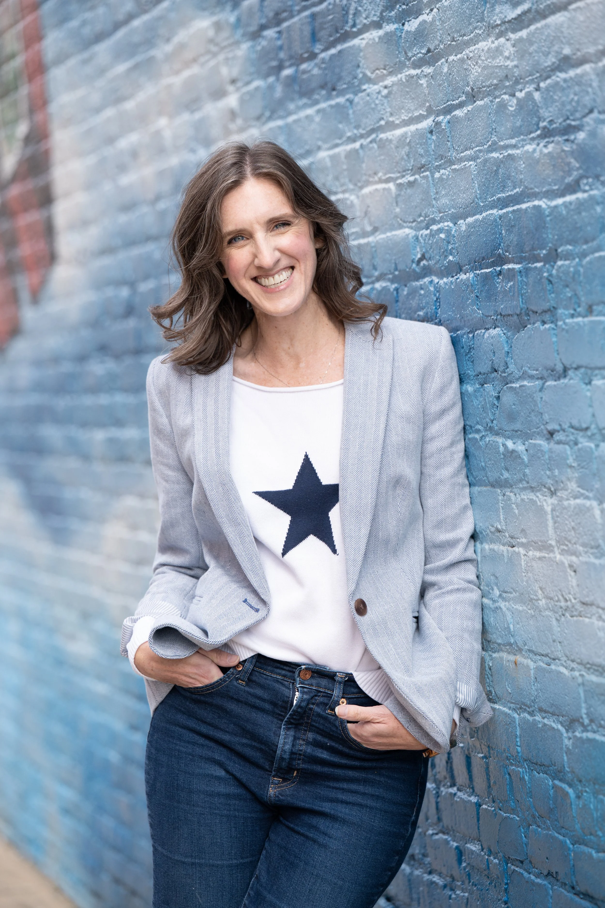 Annie Figenshu, content marketing strategist and StoryBrand Certified Guide, smiling in a blazer and star shirt against a blue brick wall.