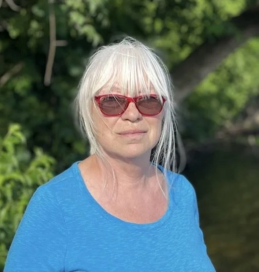 Person with white hair and red sunglasses wearing a blue shirt outdoors.