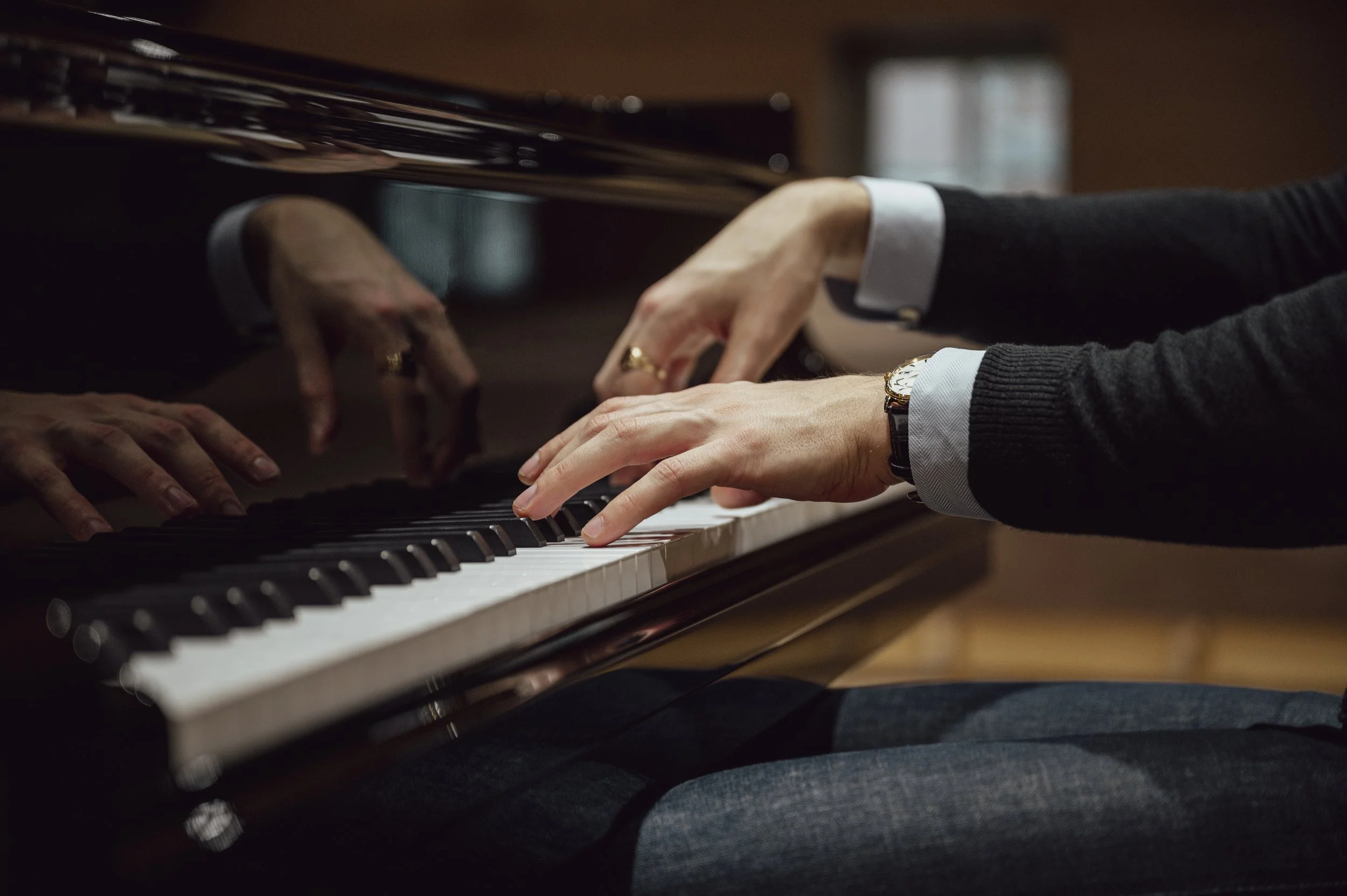 Person playing a grand piano, dressed in a dark sweater with rolled-up shirt sleeves, with a watch on their wrist.