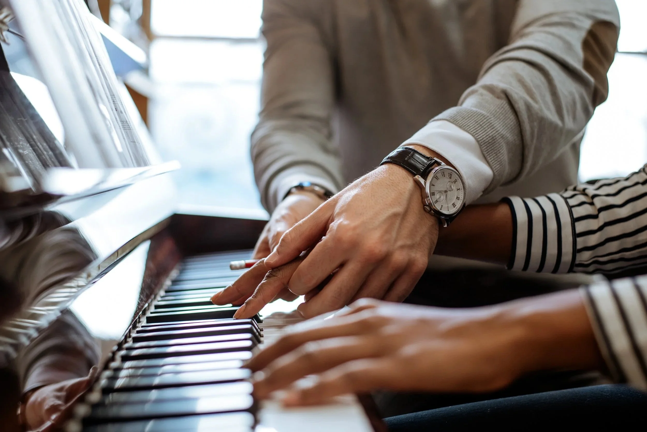 Close-up of two people playing a piano together, with one person's hand and wristwatch visible as they press piano keys.
