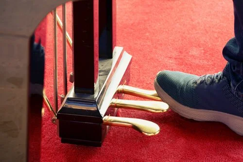 Close-up view of a piano pedal board with a person's foot pressing one pedal, on a red carpet.