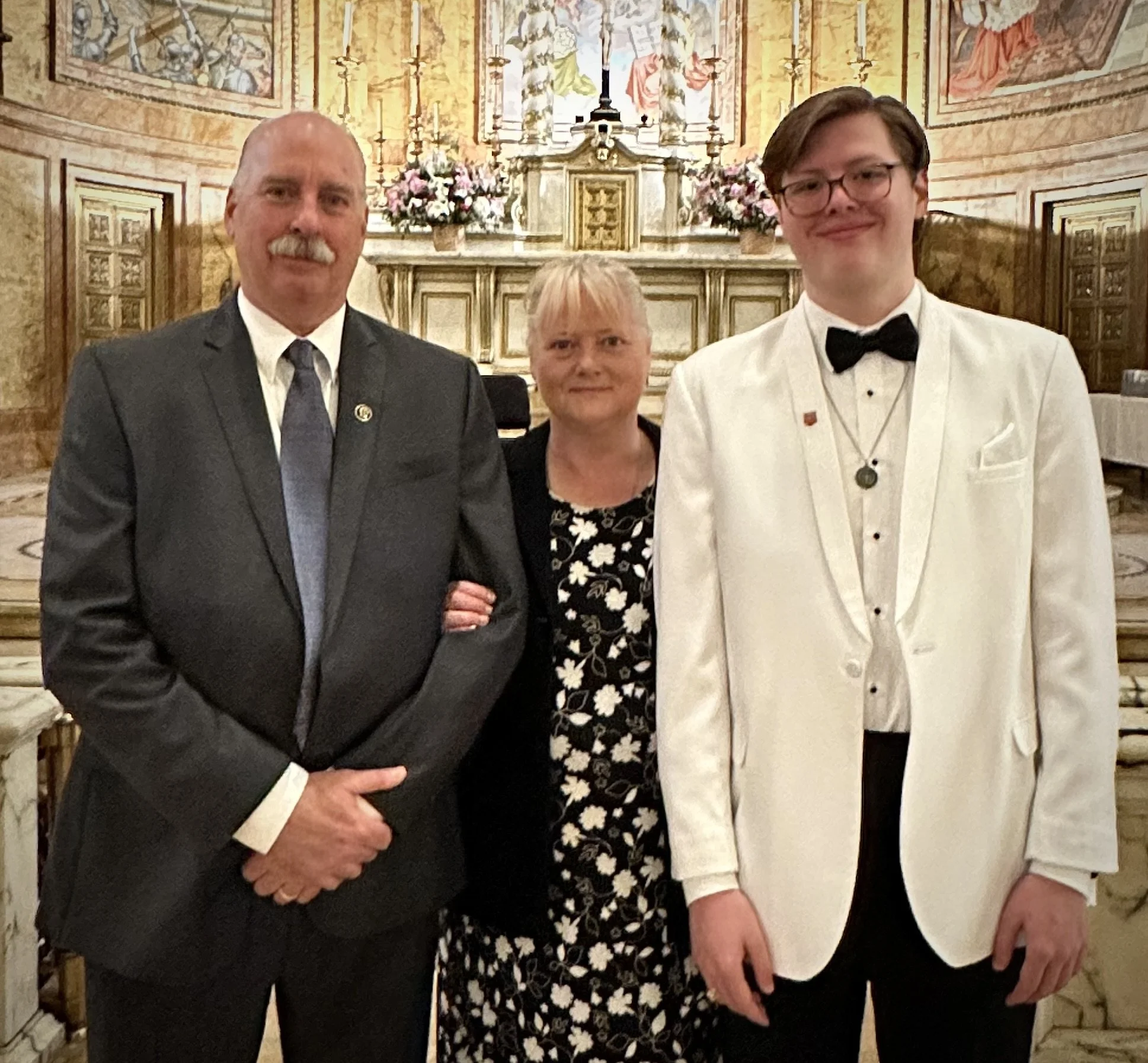 Three people posing in formal attire in front of an ornate altar with floral decorations.