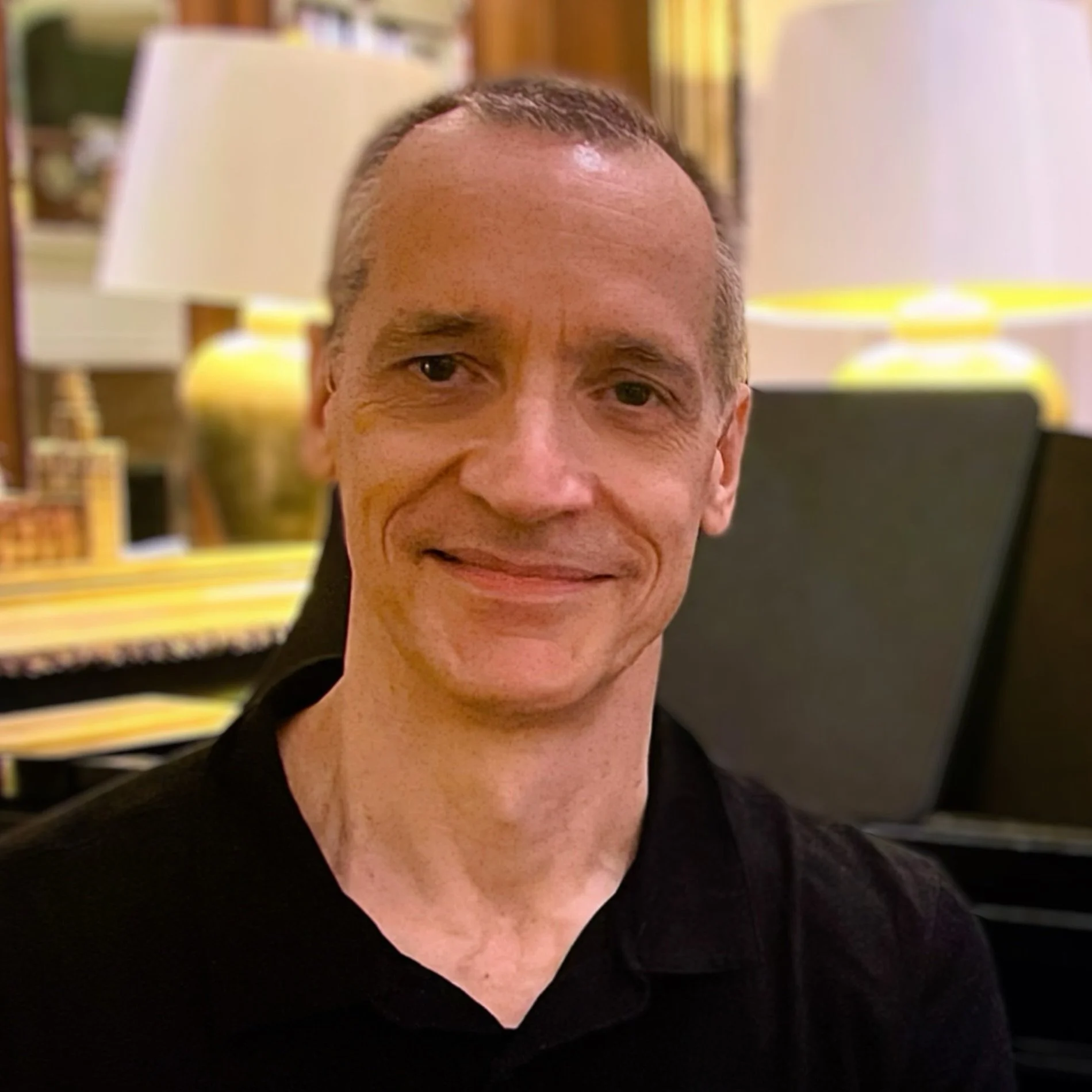 A smiling man with short hair wearing a black shirt, sitting in an indoor setting with lamps and decor in the background.