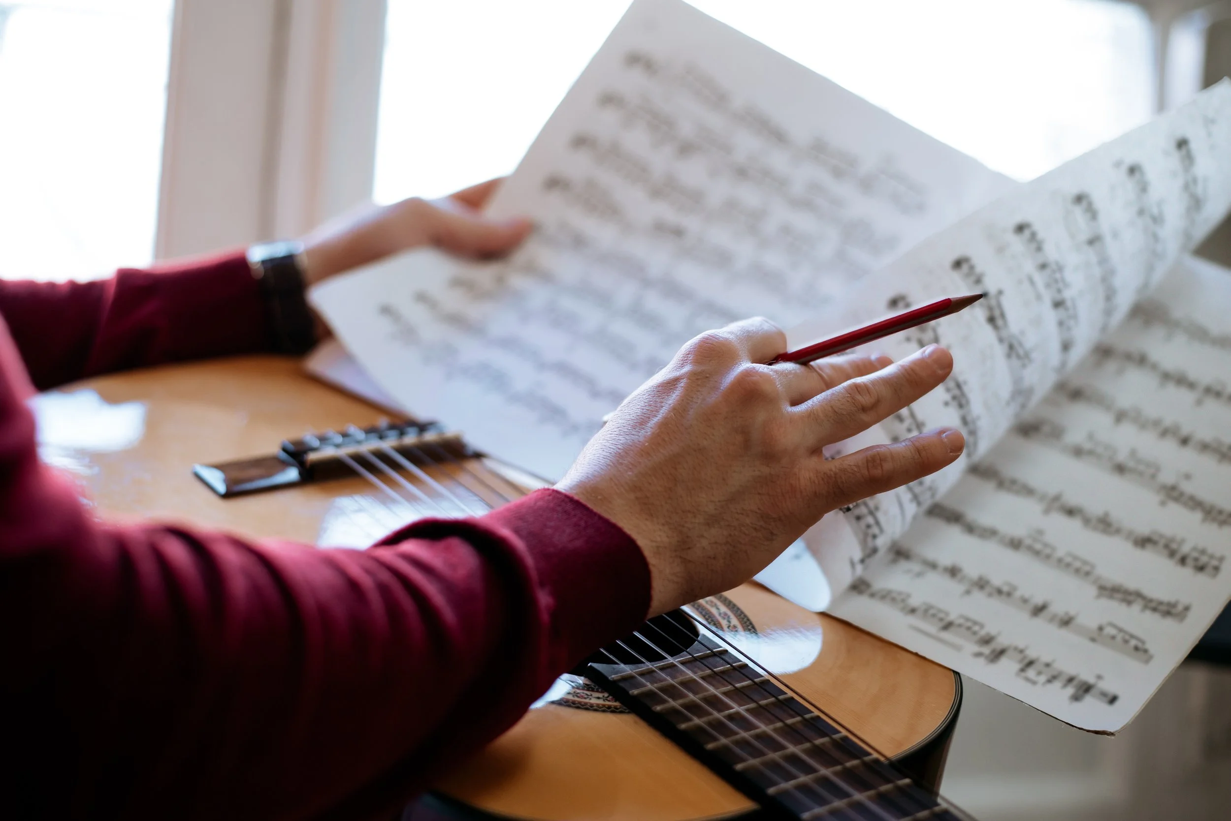 Person in a red long sleeve shirt reading sheet music while sitting at a wooden table with an acoustic guitar and a capo.