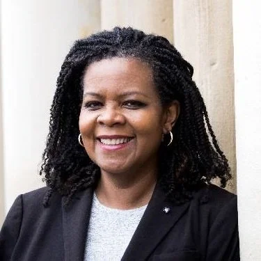 Smiling woman with braided hair, wearing a black blazer and gray top, standing outdoors against a light-colored wall.